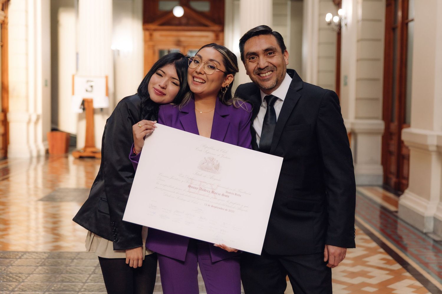 Three people posing closely together with a large white certificate in an ornate building corridor.