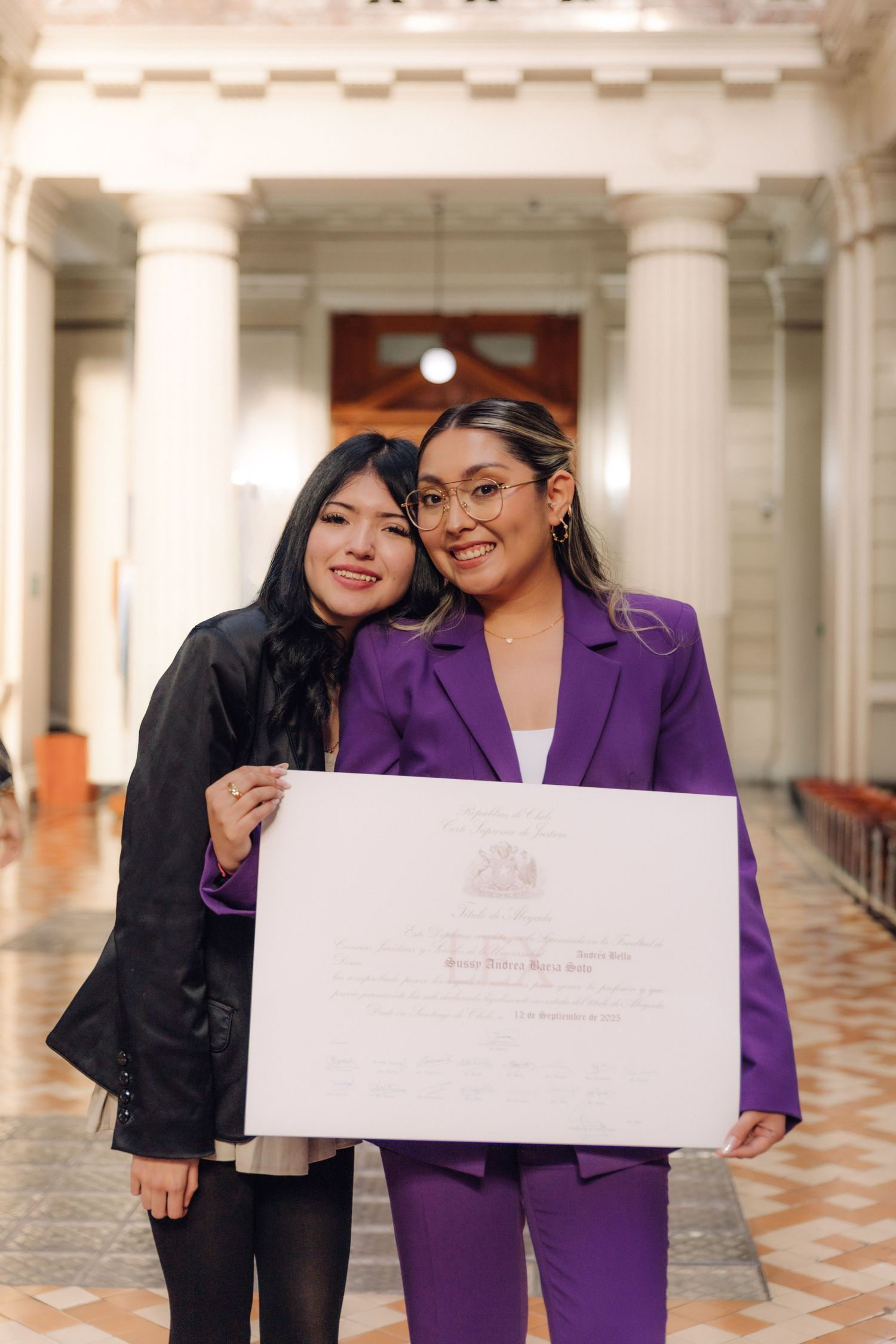 Two women pose proudly in elegant hallway holding official certificate, one wearing purple suit.