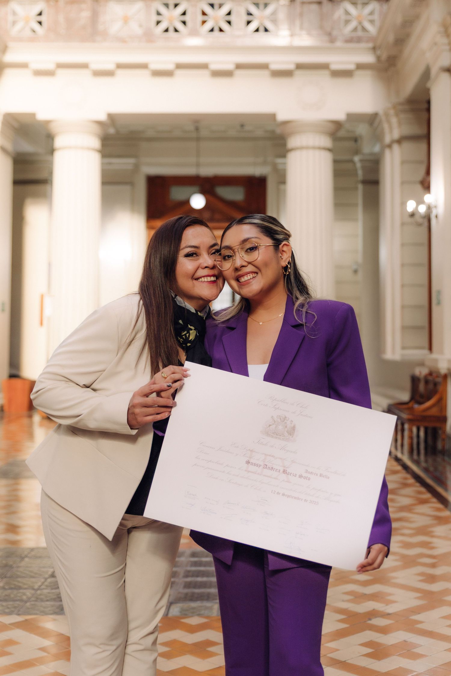 Two women in suits celebrate milestone holding official document in ornate government building.