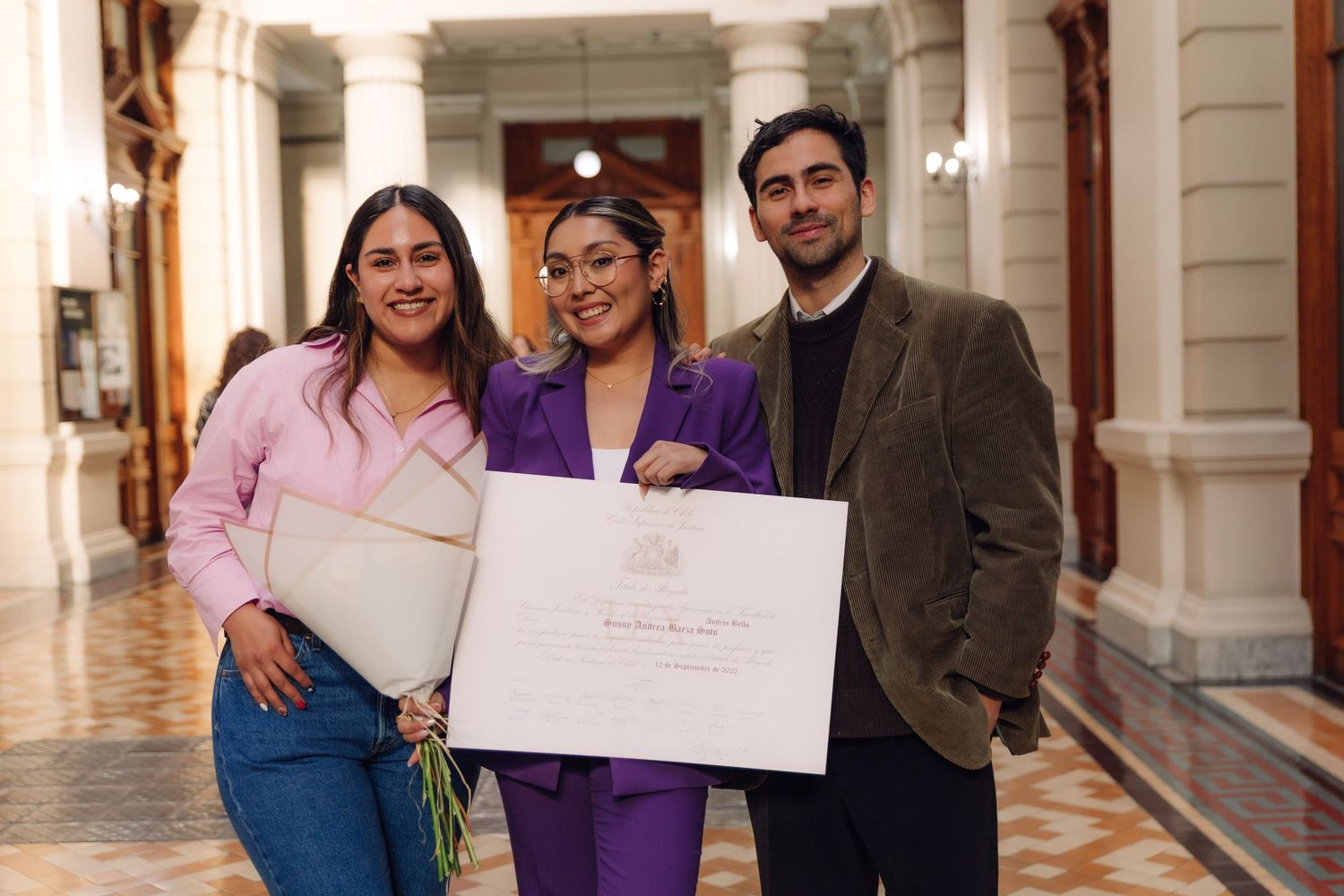 Three people smile together in hallway holding large official certificate and white folder.