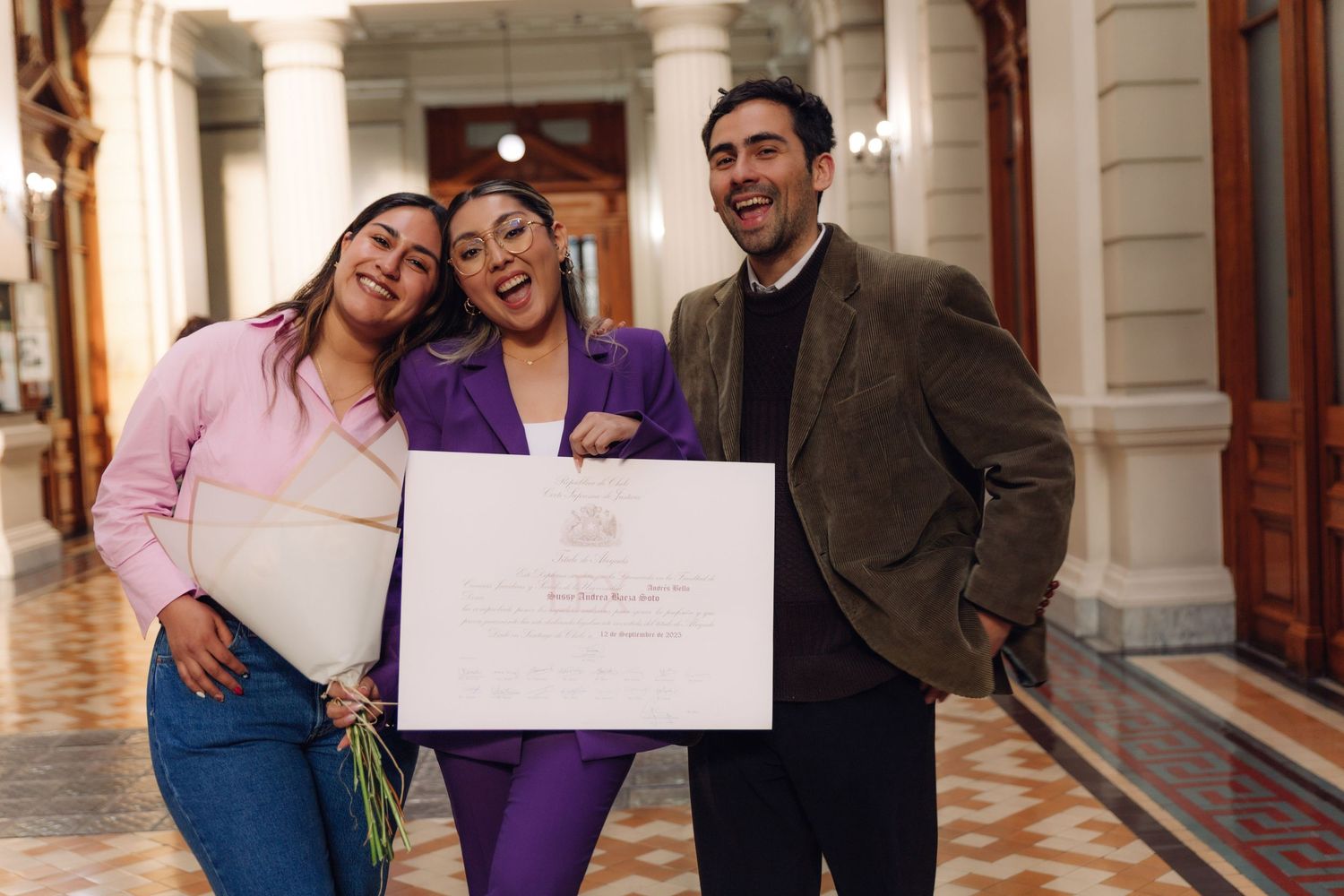 Joyful trio poses with official certificate in grand columned corridor, sharing celebration.