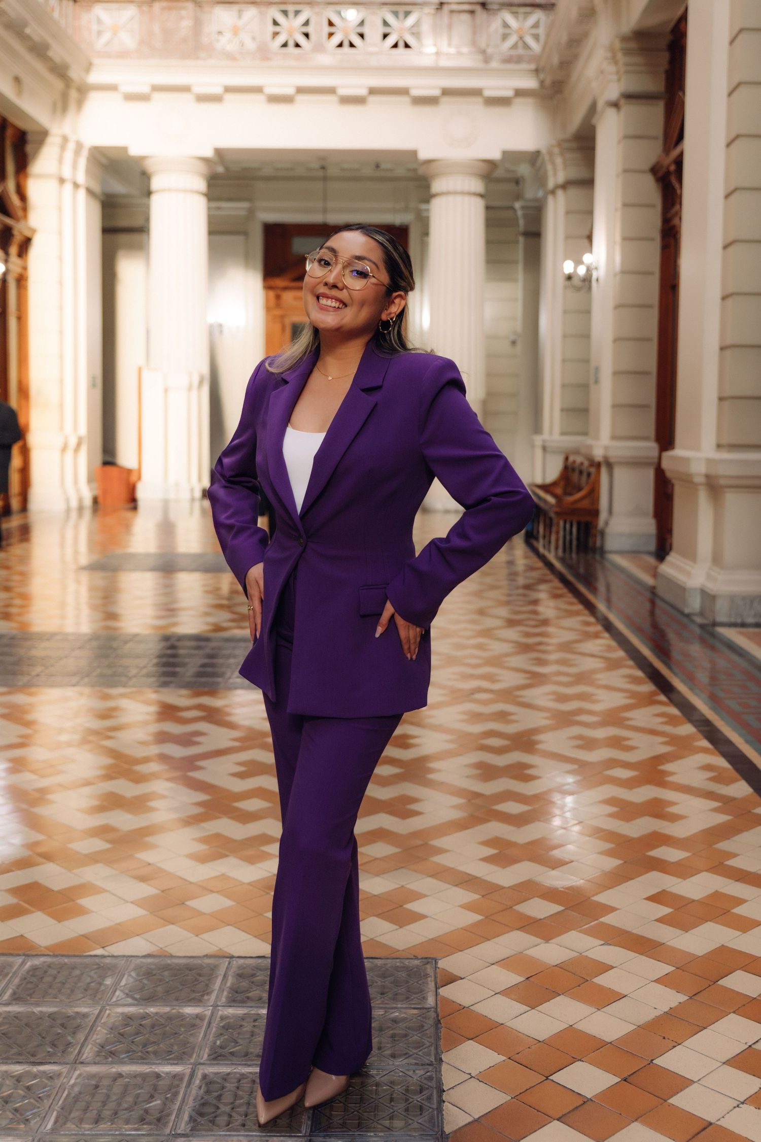 Confident woman in purple suit poses in ornate government building hallway with columns.