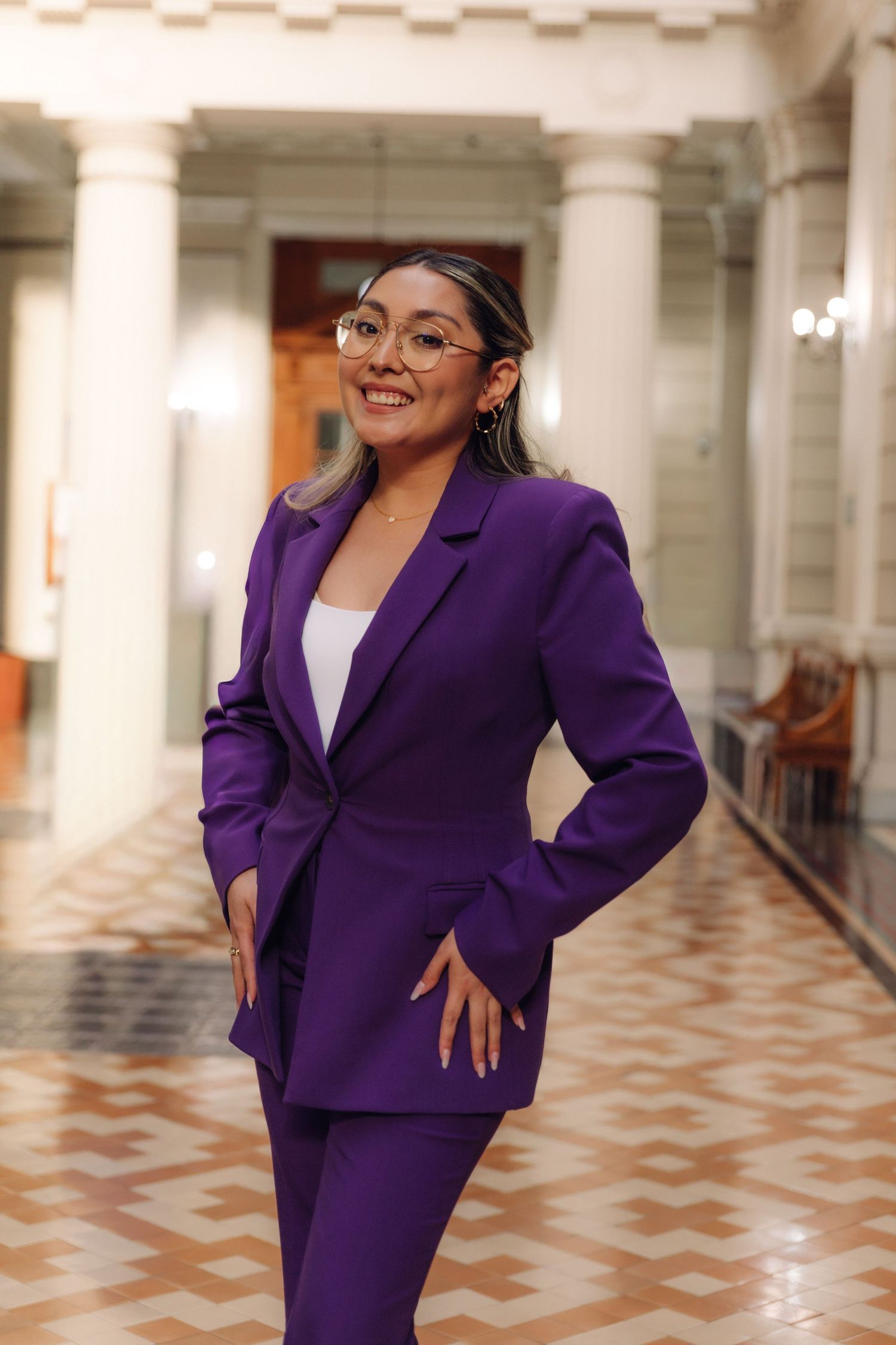 Smiling woman in purple blazer and pants stands in elegant marble-floored hallway.
