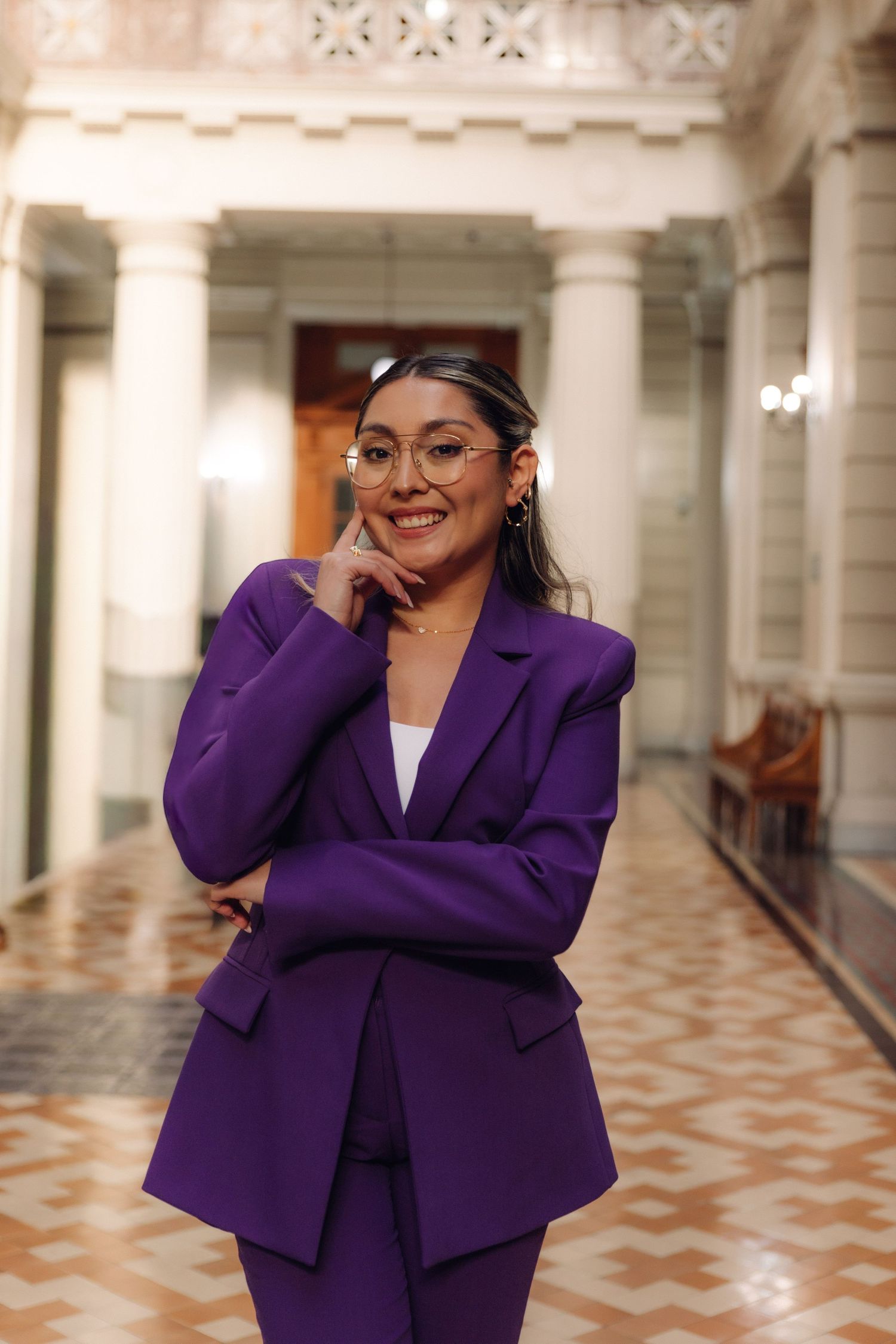 Woman in purple suit smiles thoughtfully with hand on chin in grand corridor.