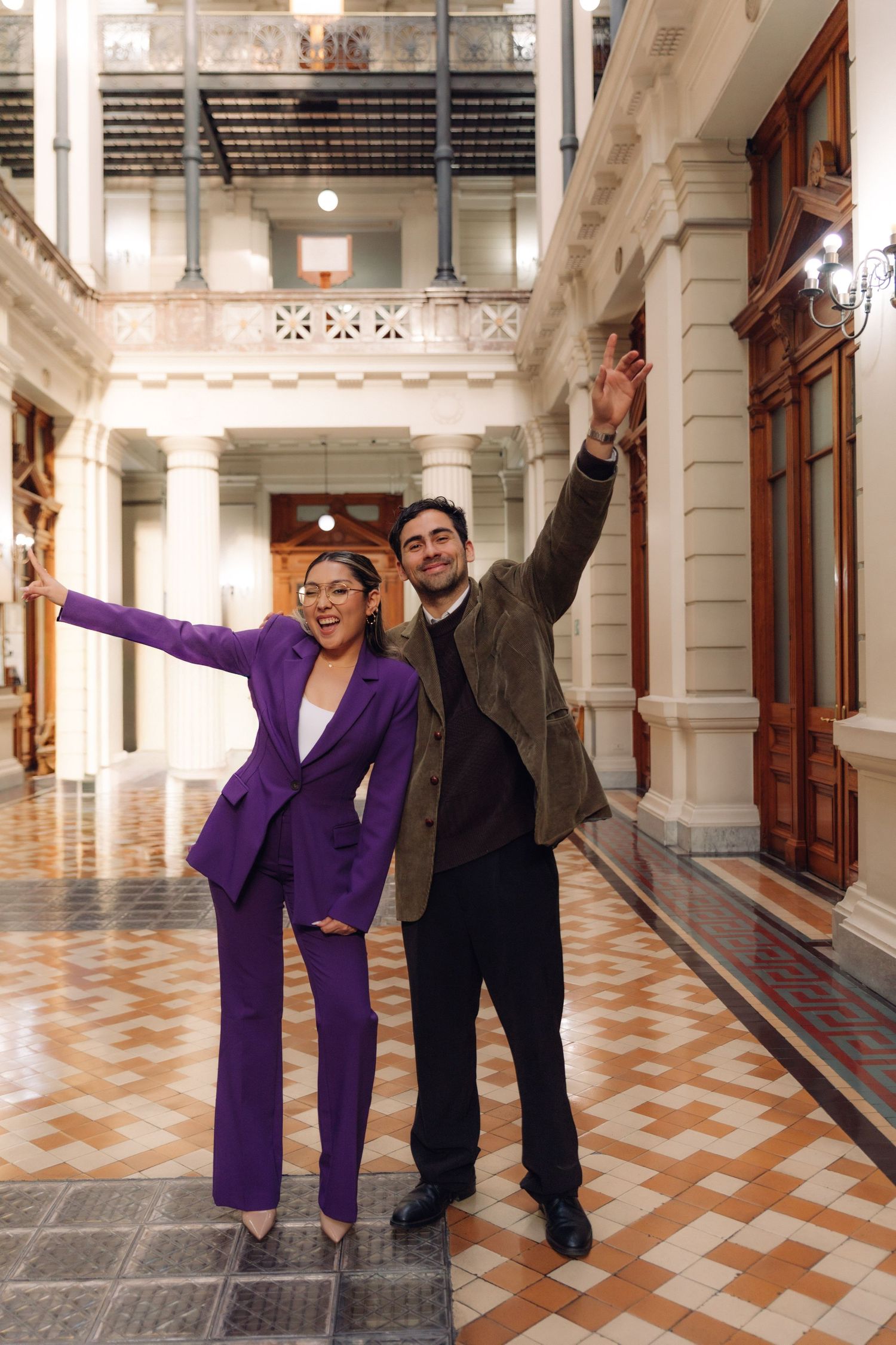 Man and woman in purple suit celebrate joyfully with raised arms in ornate hallway.