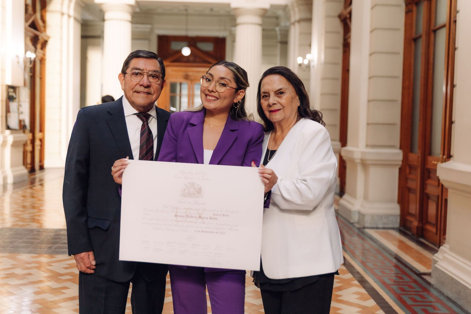 Three people proudly display official certificate in elegant government building hallway.