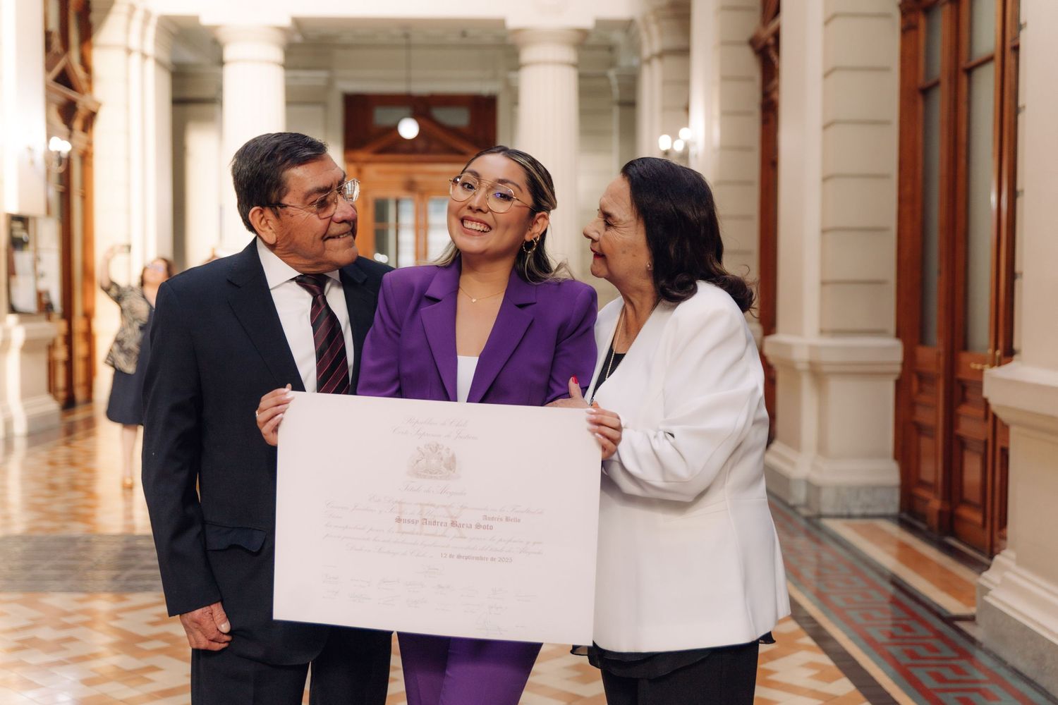 Woman in purple suit holds diploma with proud parents in elegant hallway.