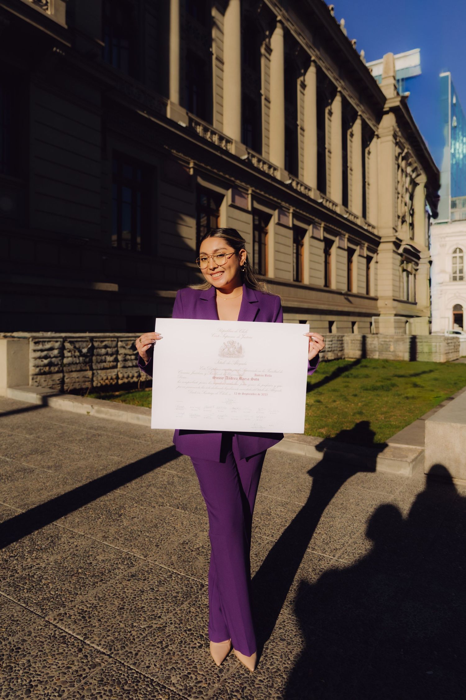 Woman in purple suit holds official certificate outside historic government building in sunlight.