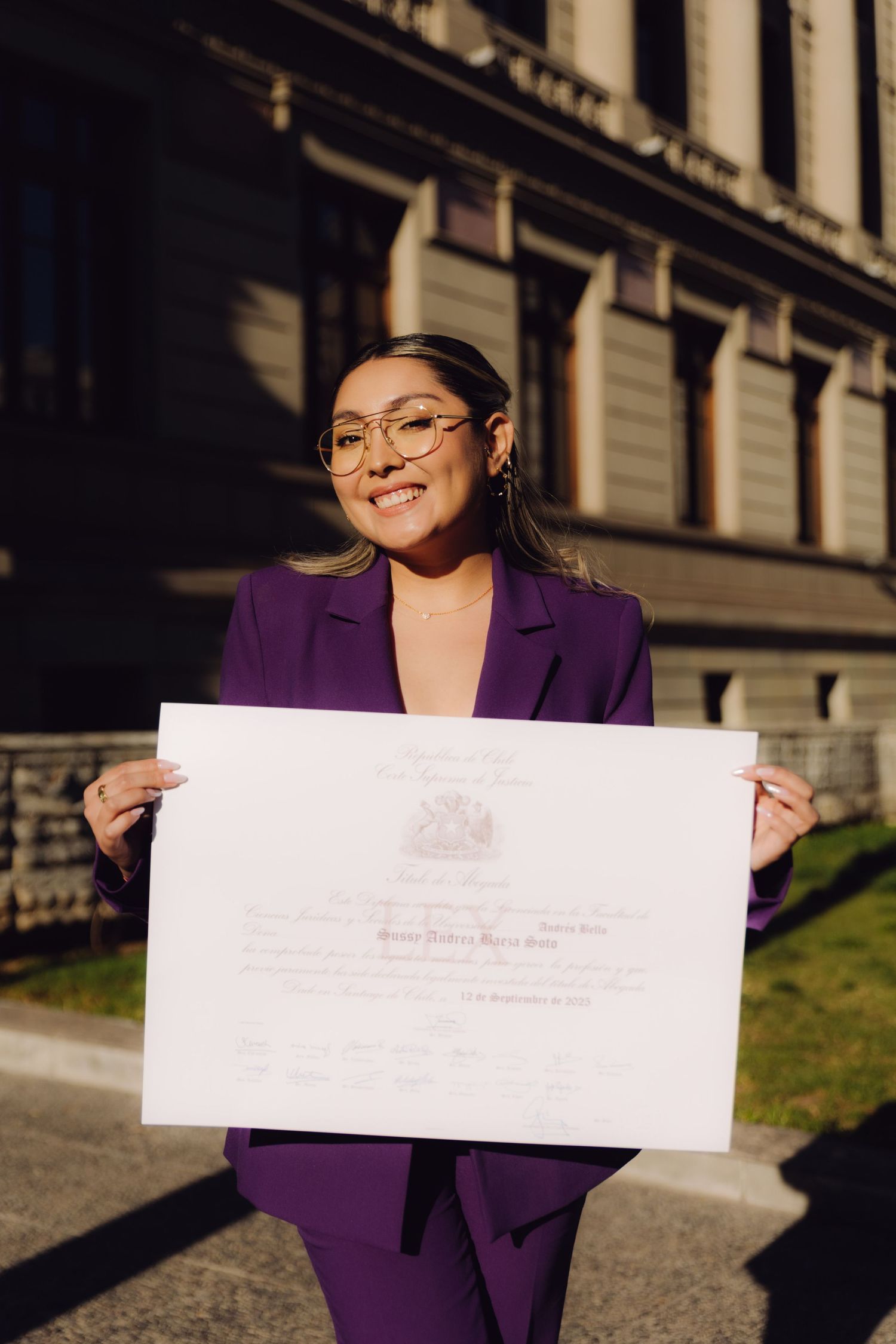 Young woman in purple suit proudly holds official diploma outside historic building.