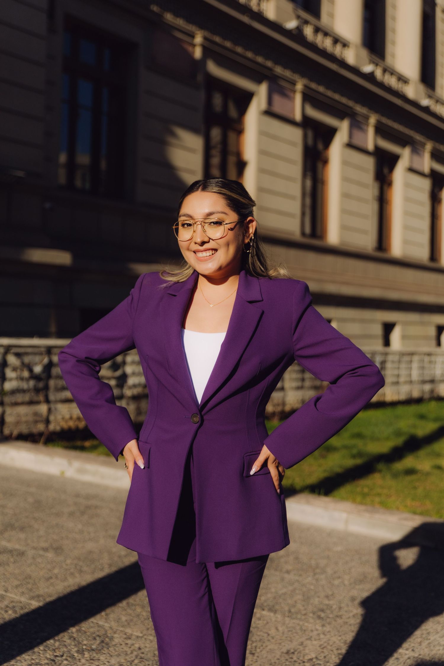 Smiling woman in purple suit with glasses poses outside historic stone building.