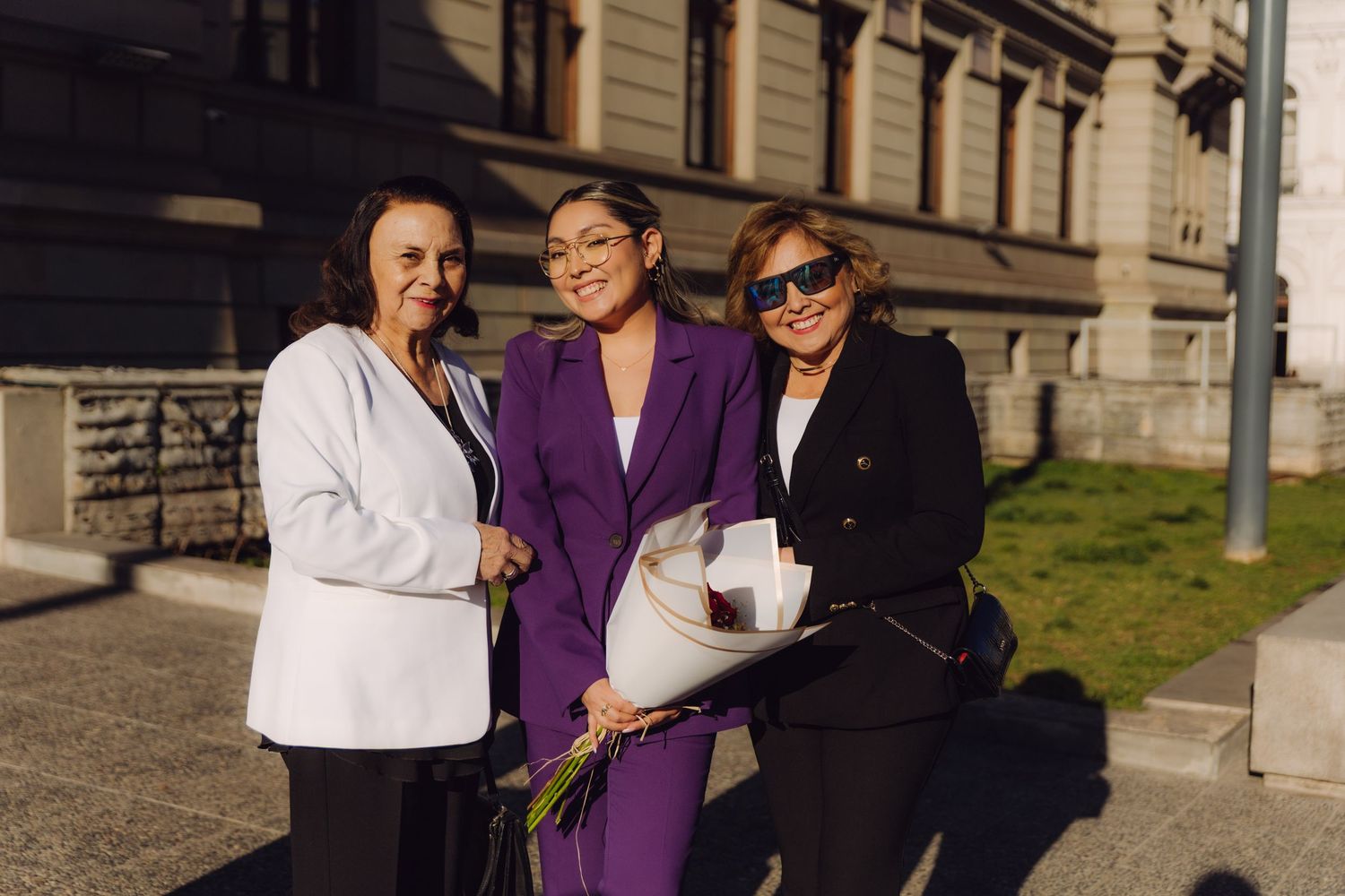 Three women smile together outside sunlit historic building, holding flowers and documents.