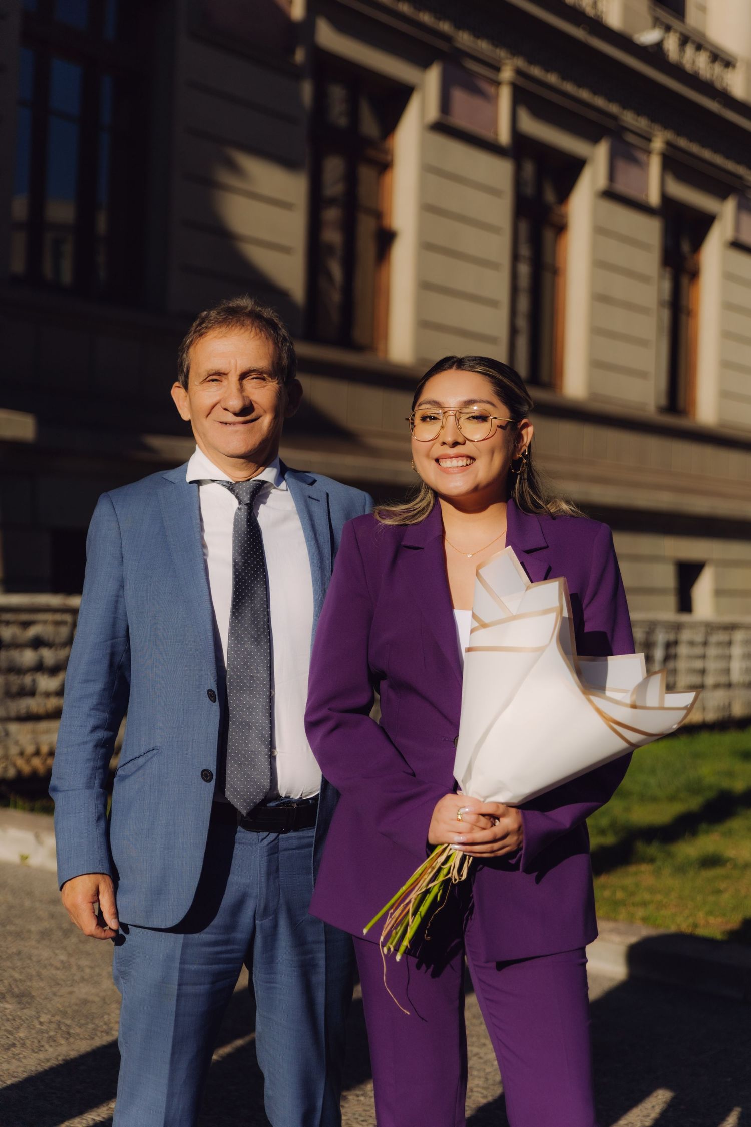 Man in blue suit and woman in purple suit pose with bouquet outside building.