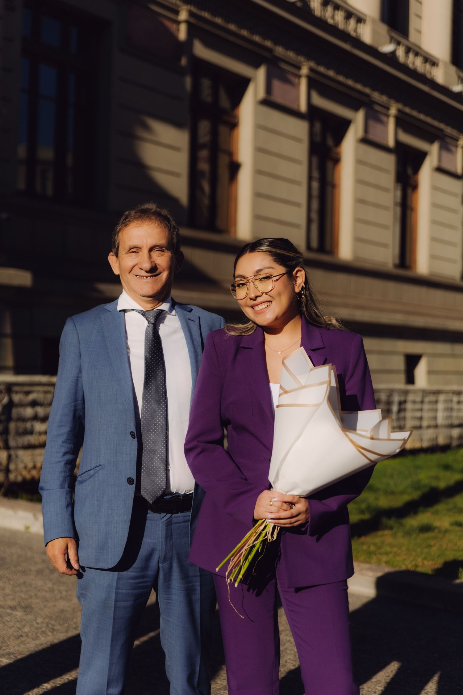 Smiling man in blue suit and woman in purple suit pose with white bouquet outdoors.