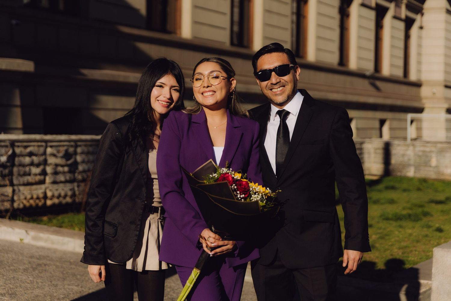 Three people smile together outdoors, woman in purple suit holds colorful flower bouquet.