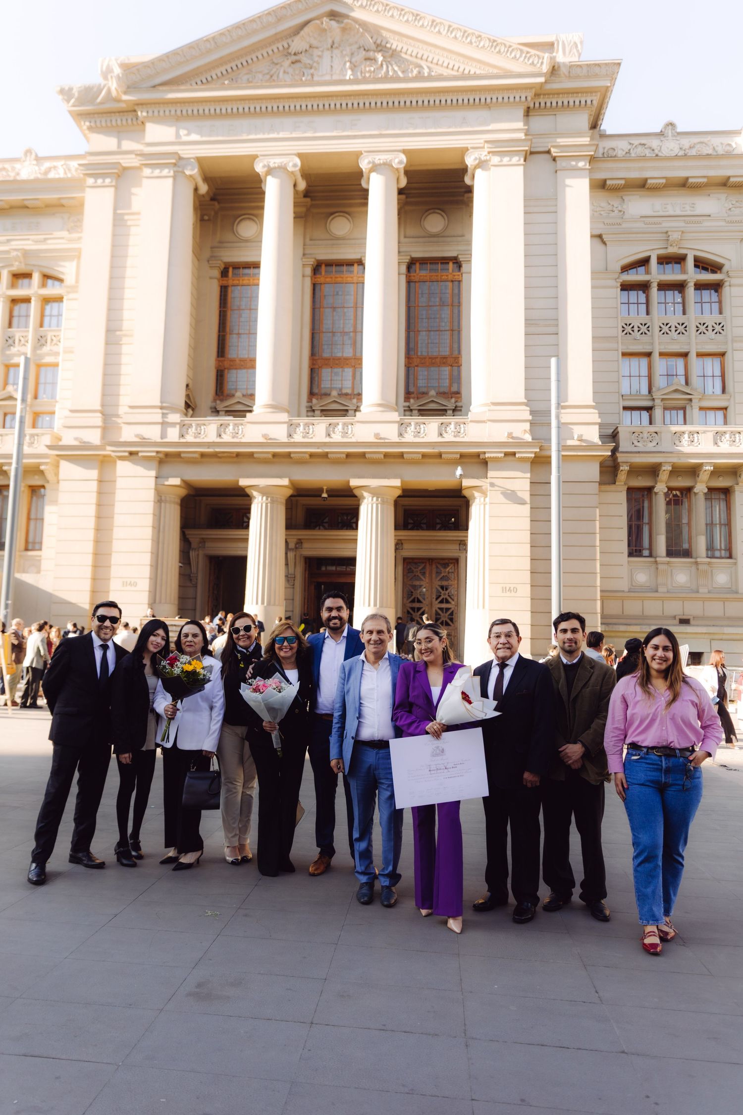 Group of professionals pose before grand columned building with diploma and flowers.