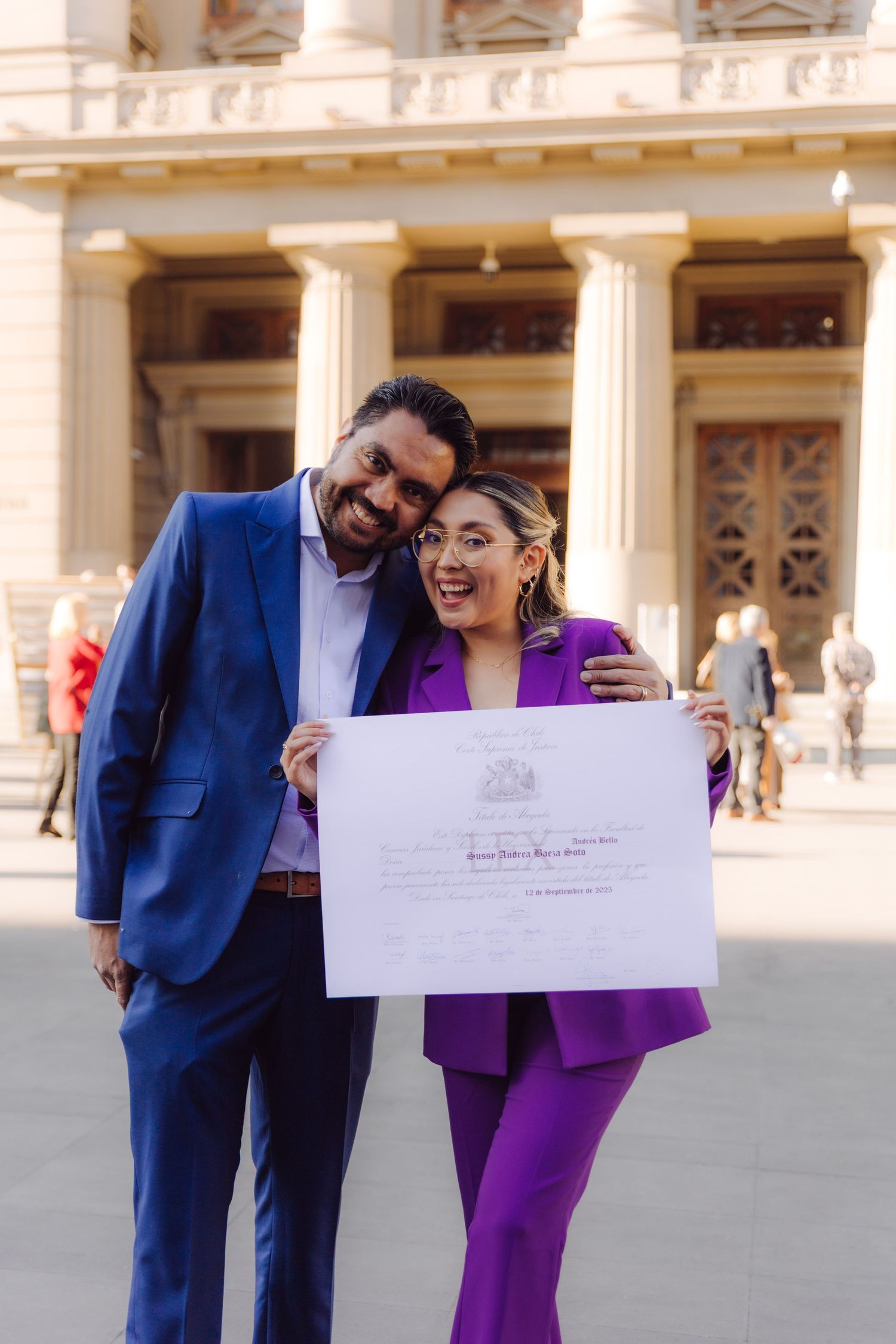 Couple in blue suit and purple pantsuit proudly holding official certificate outside neoclassical building.