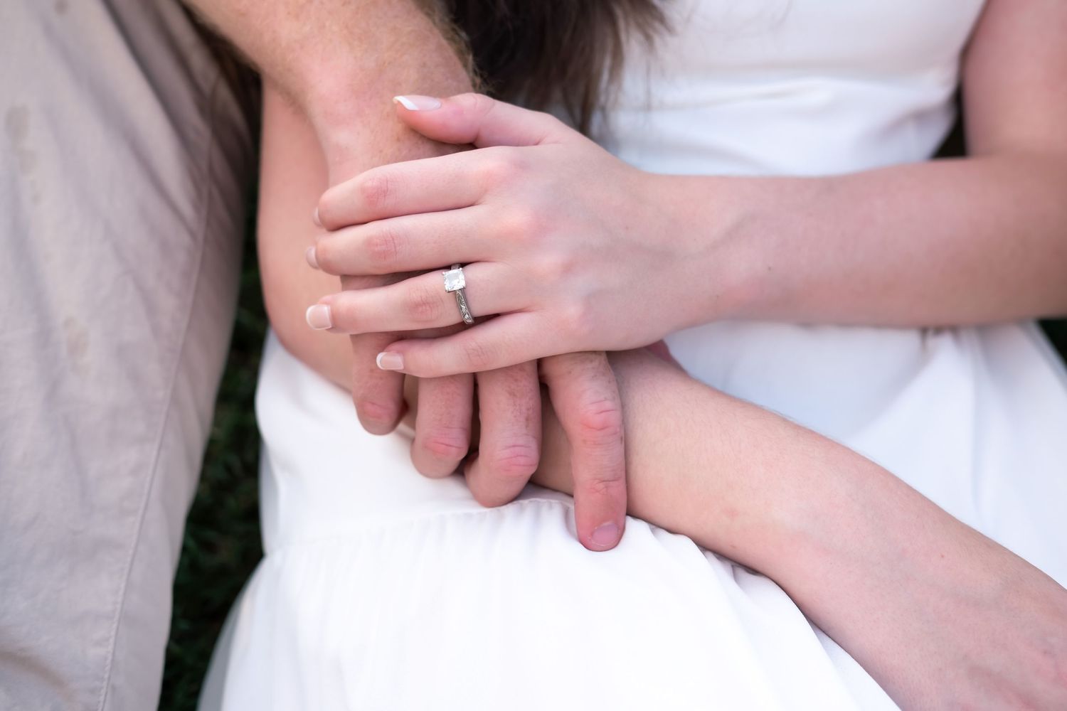 Close-up of a couple holding hands, woman wearing a diamond engagement ring, dressed in white wedding attire.