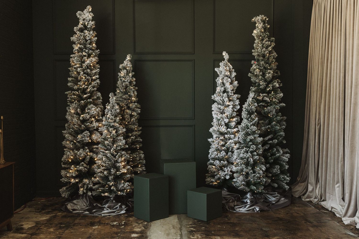 Three flocked Christmas trees with warm lights beside green gift boxes against a dark green paneled wall.