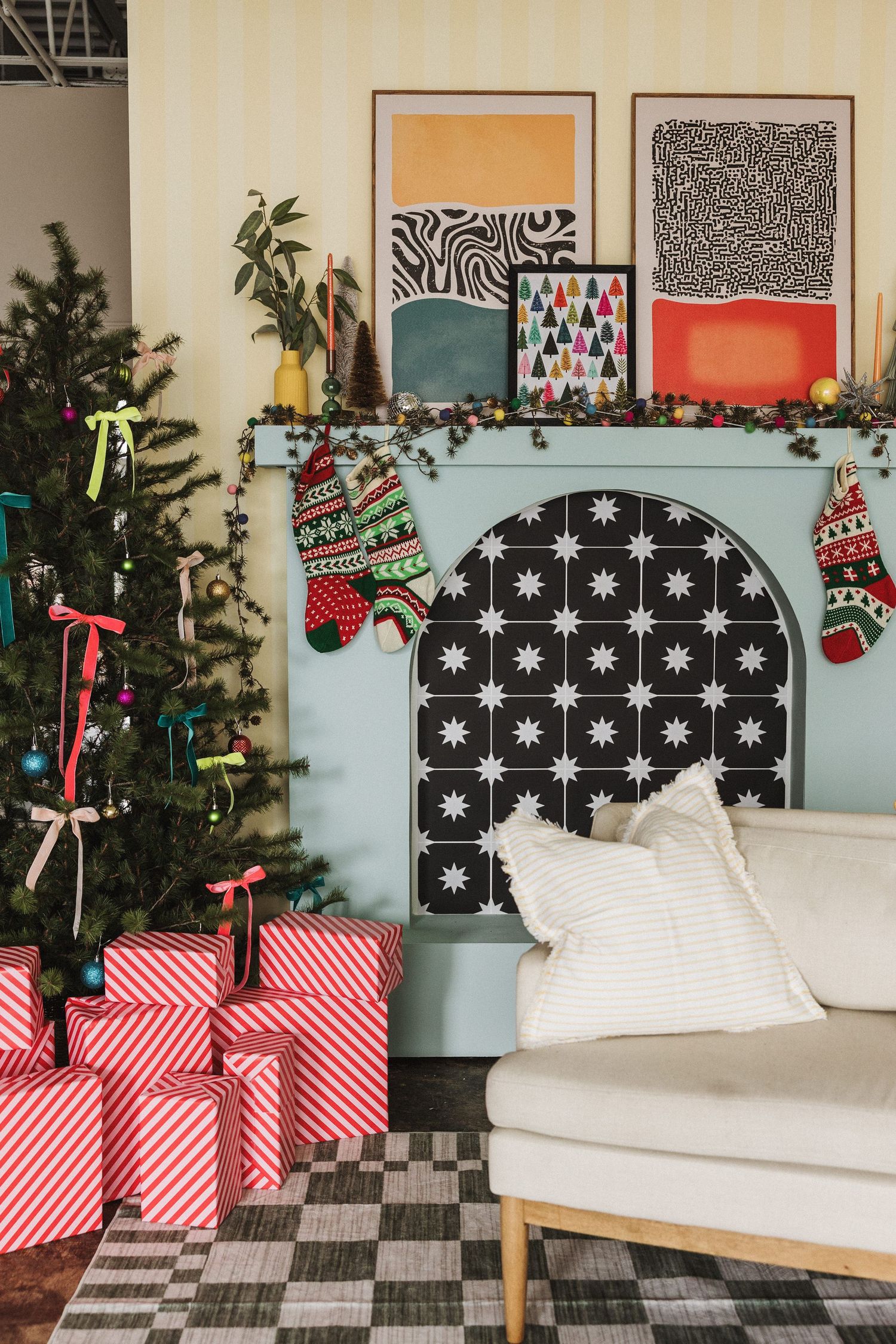 Festive living room with decorated Christmas tree, wrapped gifts, stockings on a tiled fireplace, and colorful wall art.