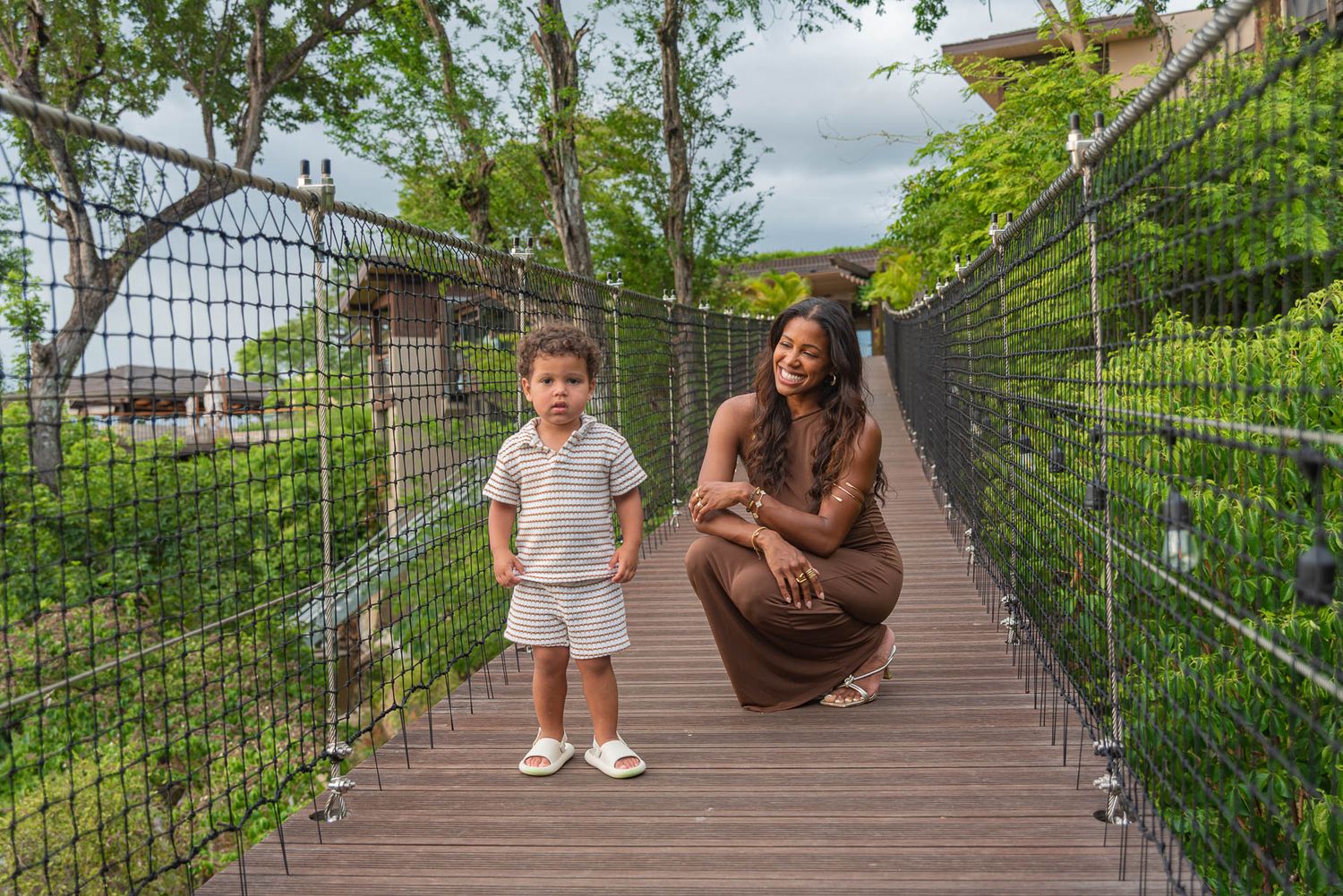 A woman crouches beside a young child on a wooden suspension bridge surrounded by lush green trees.