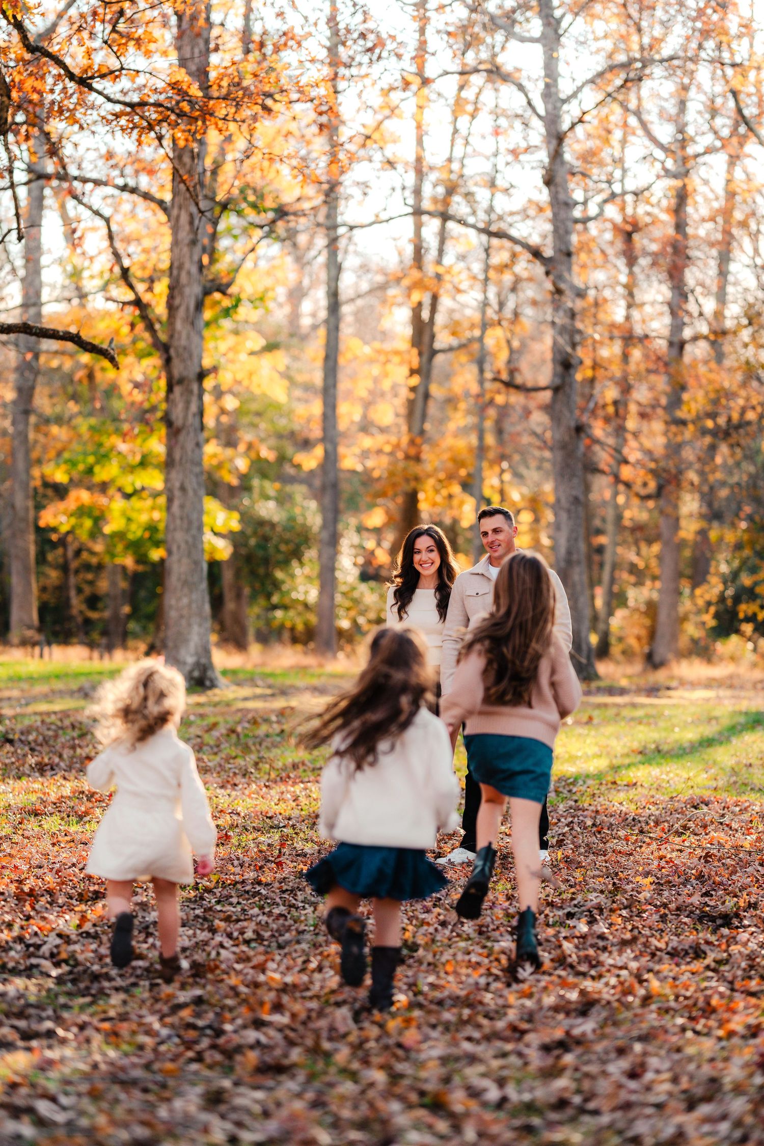 Family playing in autumn forest with fallen leaves, two girls running toward smiling parents in warm fall colors.