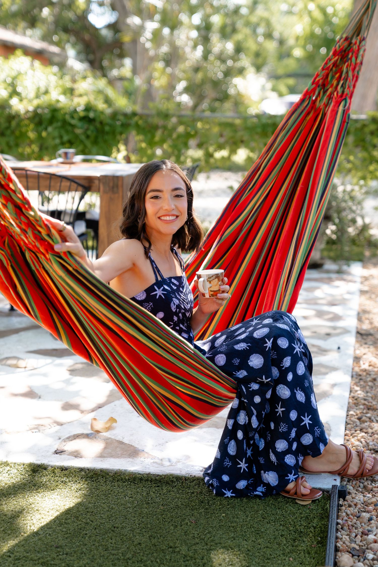Smiling woman relaxing in a colorful striped hammock outdoors, wearing a navy floral jumpsuit and holding a cup.