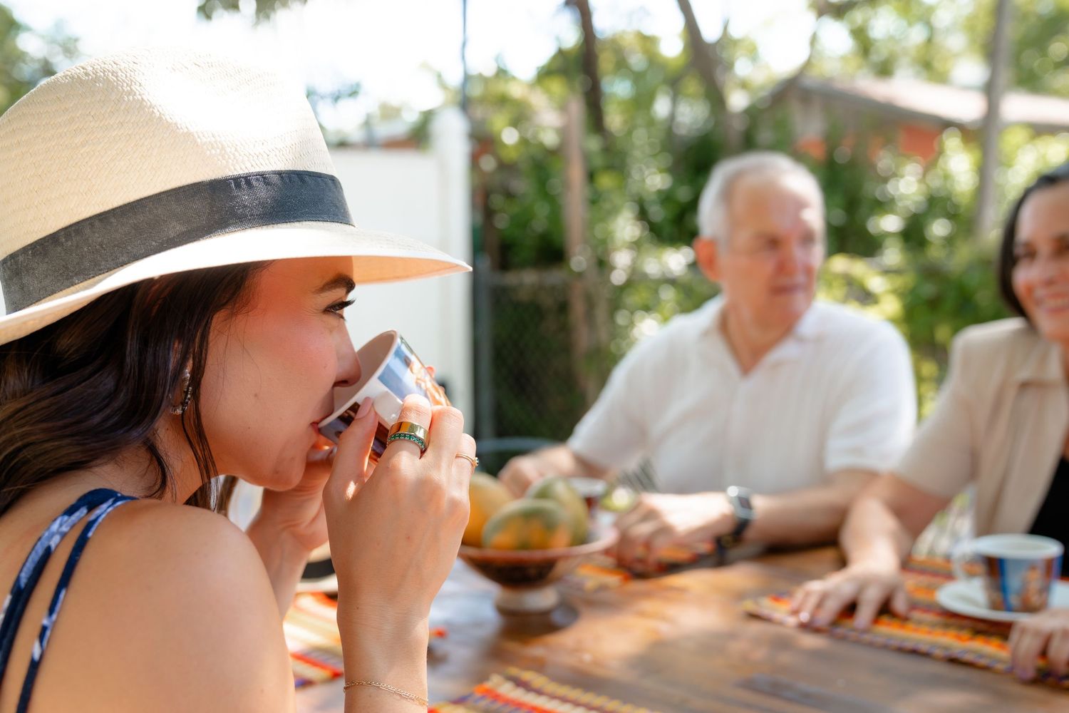 Woman in sun hat drinking outdoors at a wooden table with two people dining together in a sunny garden.
