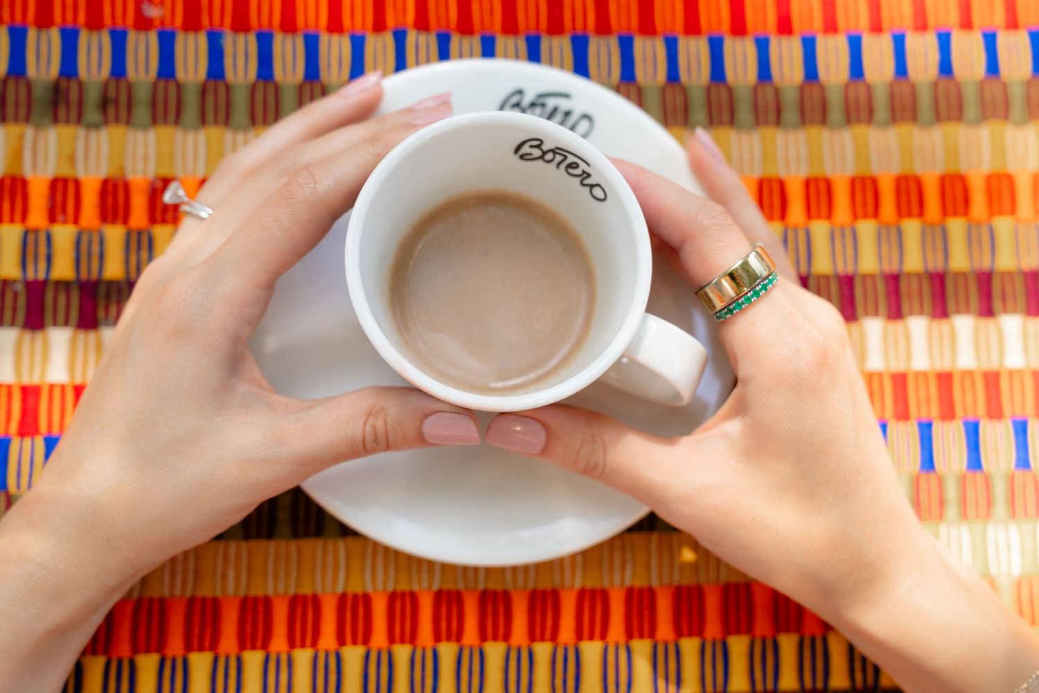 Hands holding a white coffee cup with saucer on a colorful striped placemat, with a gold ring visible.