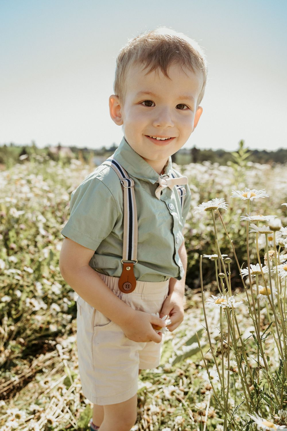Smiling young boy in suspenders and sage shirt standing in a sunny daisy field.