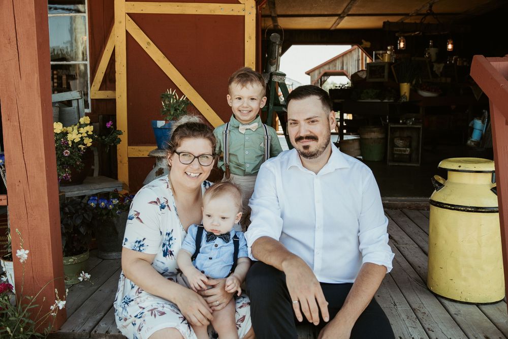 A happy family of four poses on a rustic farm porch, with two young boys and their smiling parents in casual attire.