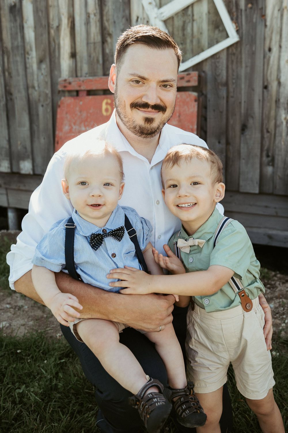 A father crouches outdoors holding two young boys in bow ties and suspenders, smiling together near a rustic wooden barn.