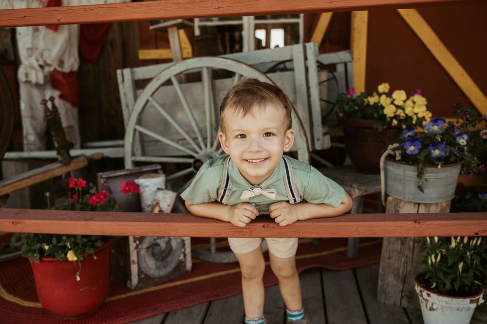 A smiling toddler boy leans on a wooden railing surrounded by colorful flowers and a vintage wagon wheel.