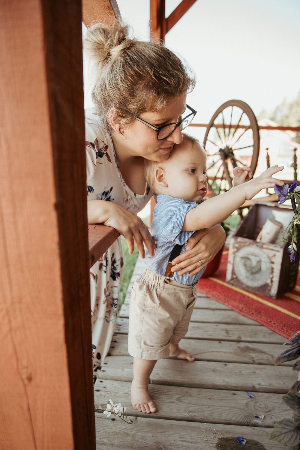 A mother with glasses kisses her toddler on a wooden porch, as the baby reaches toward purple flowers.