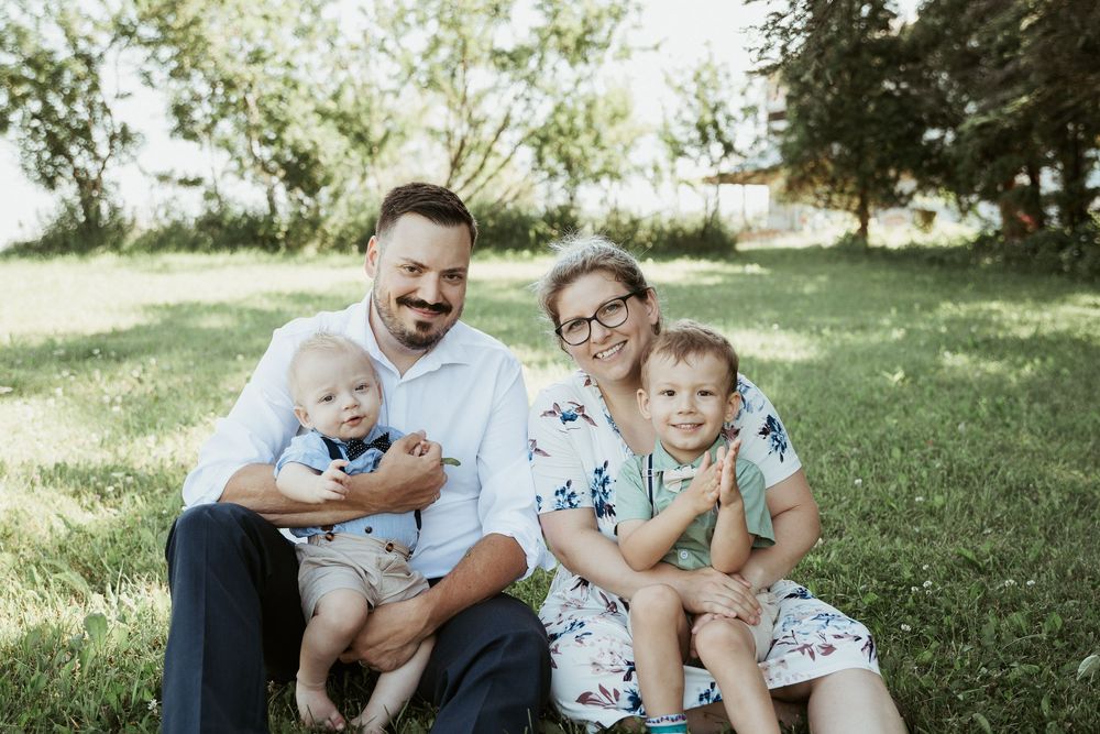 Happy family of four sitting on green grass outdoors, parents holding two young toddler boys, smiling in a sunny park.