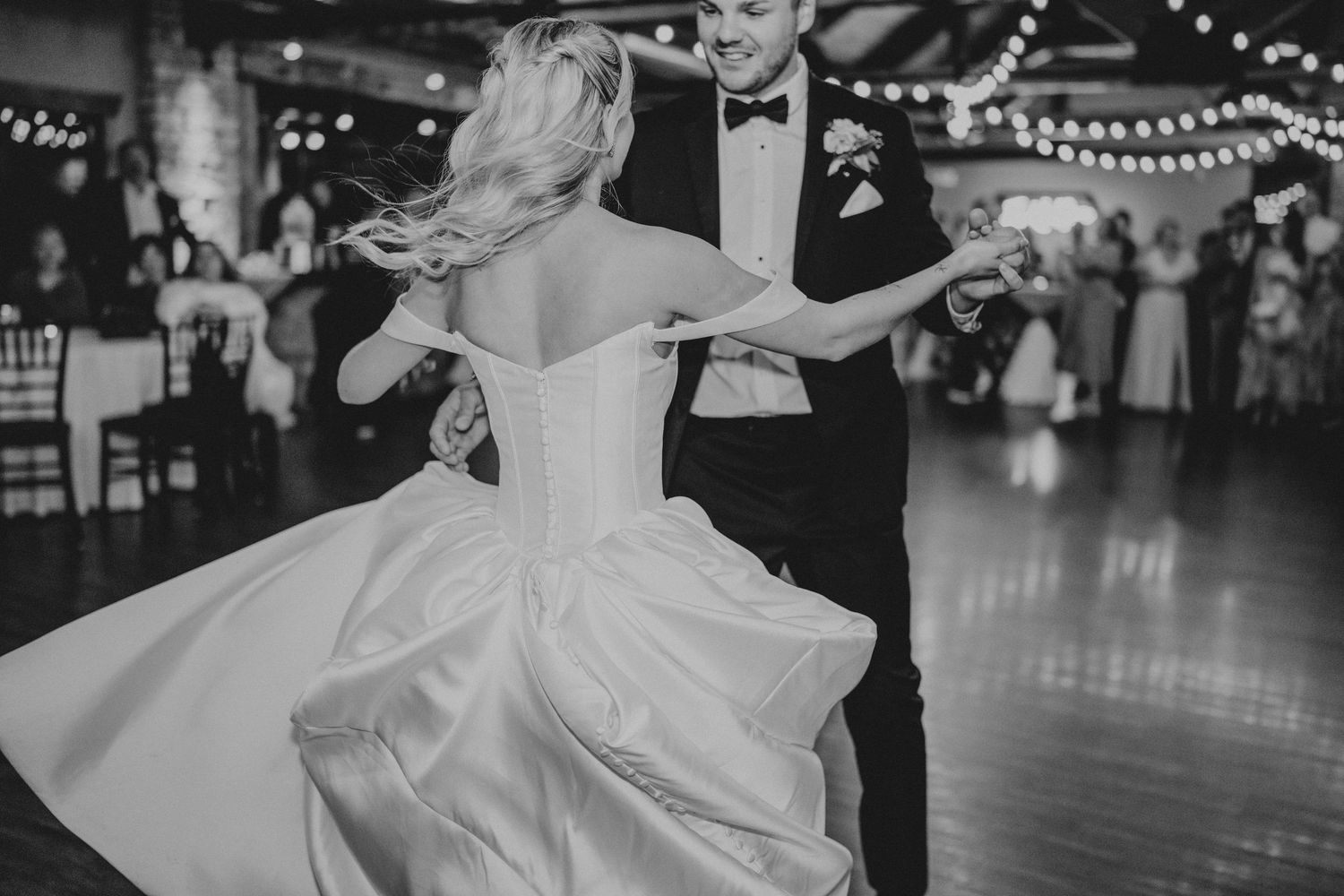 Black and white photo of bride and groom sharing first dance at elegant wedding reception with string lights.