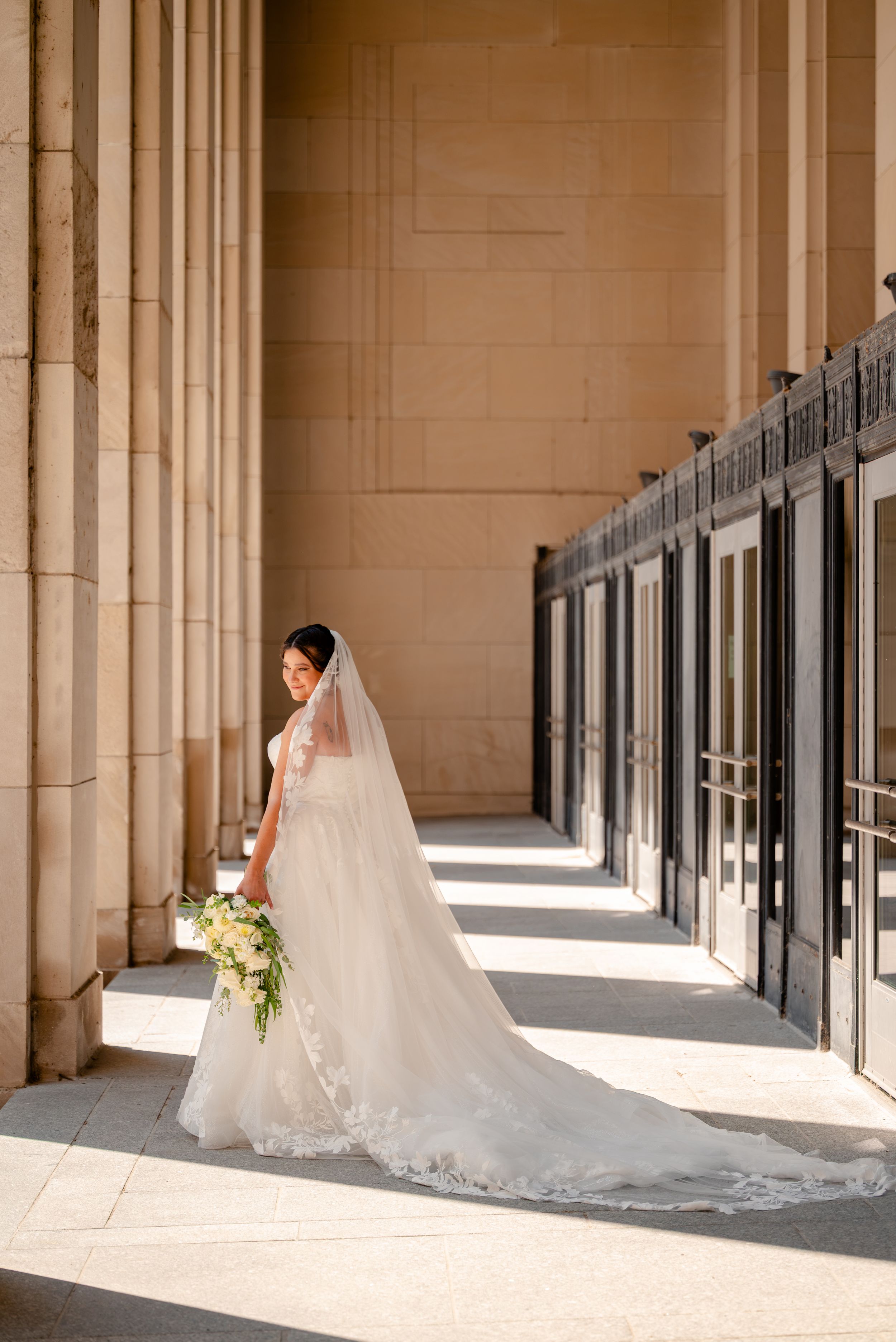 Bride in white lace gown and veil holding bouquet beside tall stone columns in elegant venue.