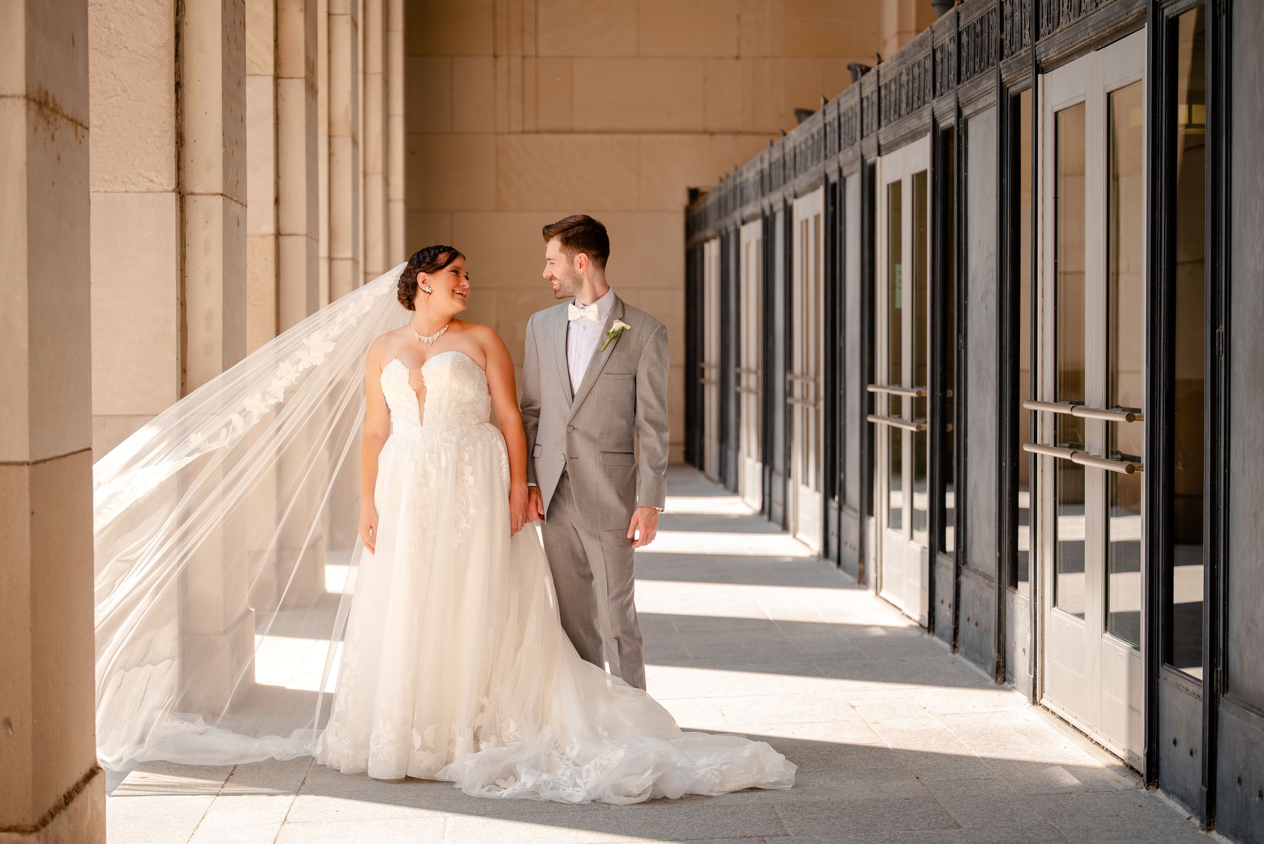 Bride in white gown and groom in gray suit hold hands outside elegant columned building on their wedding day.