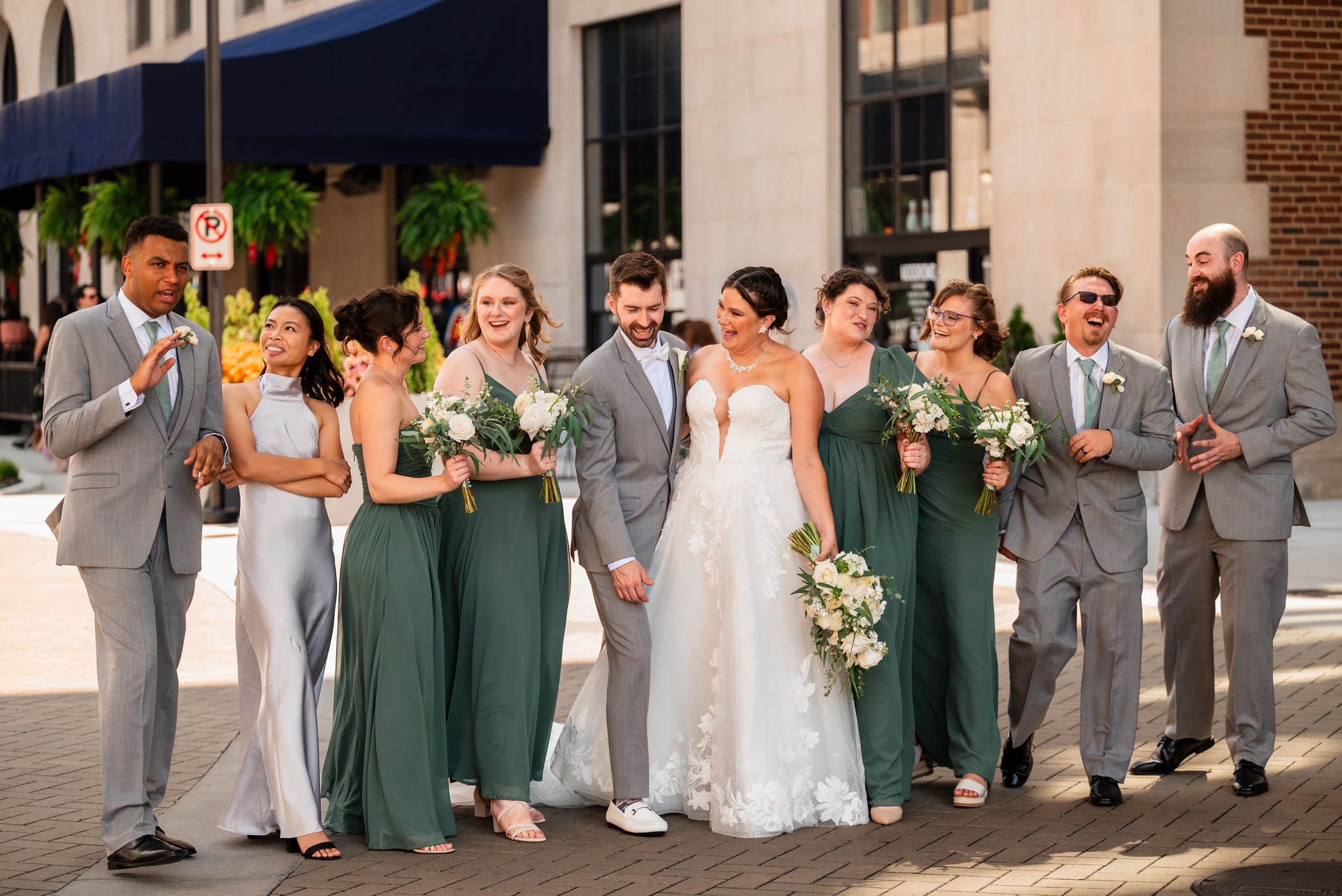 Wedding party walks outdoors, bride and groom centered, bridesmaids in green dresses, groomsmen in gray suits, holding bouquets.