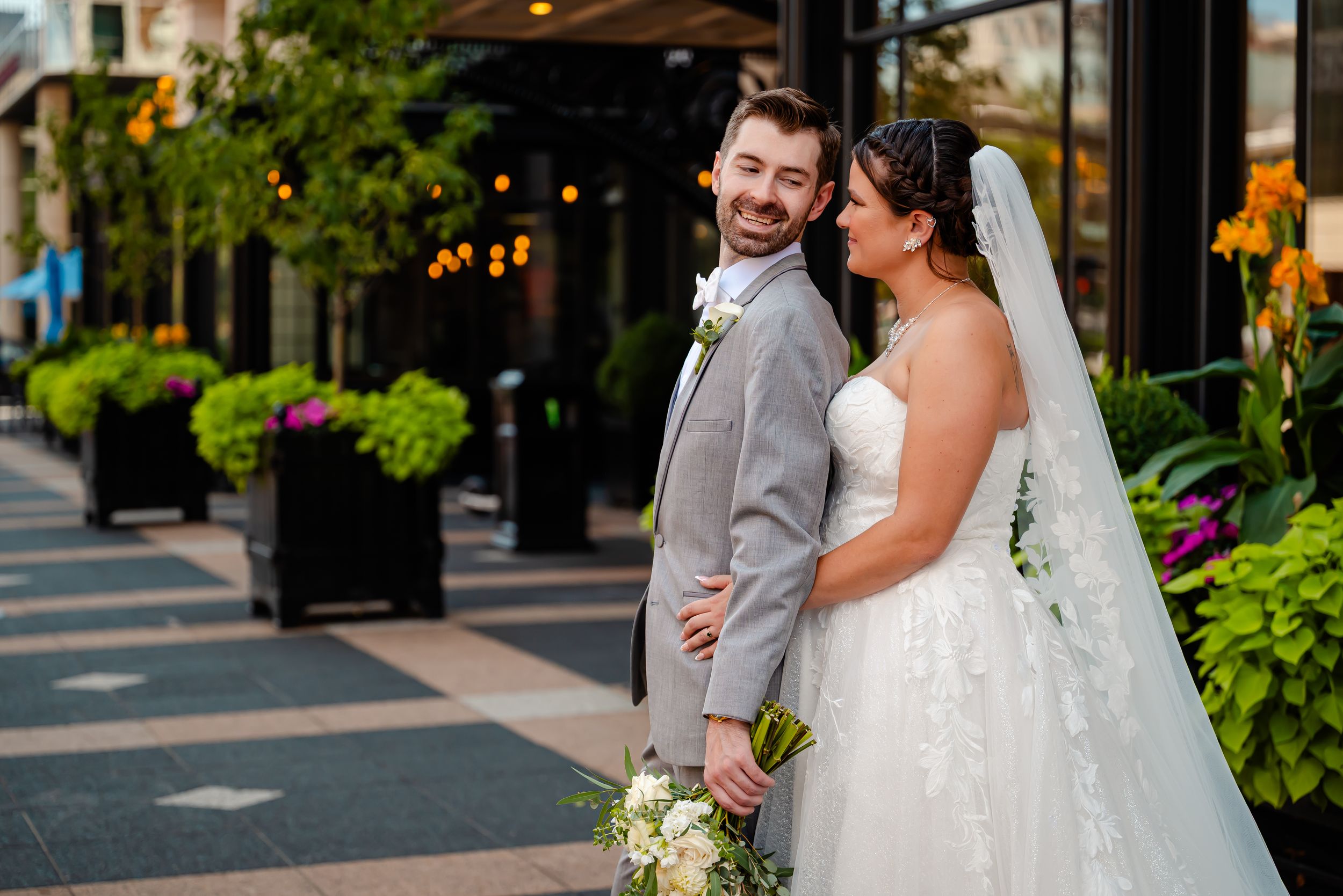 Bride and groom pose on an outdoor urban walkway, she holds white flowers, both smiling in wedding attire.