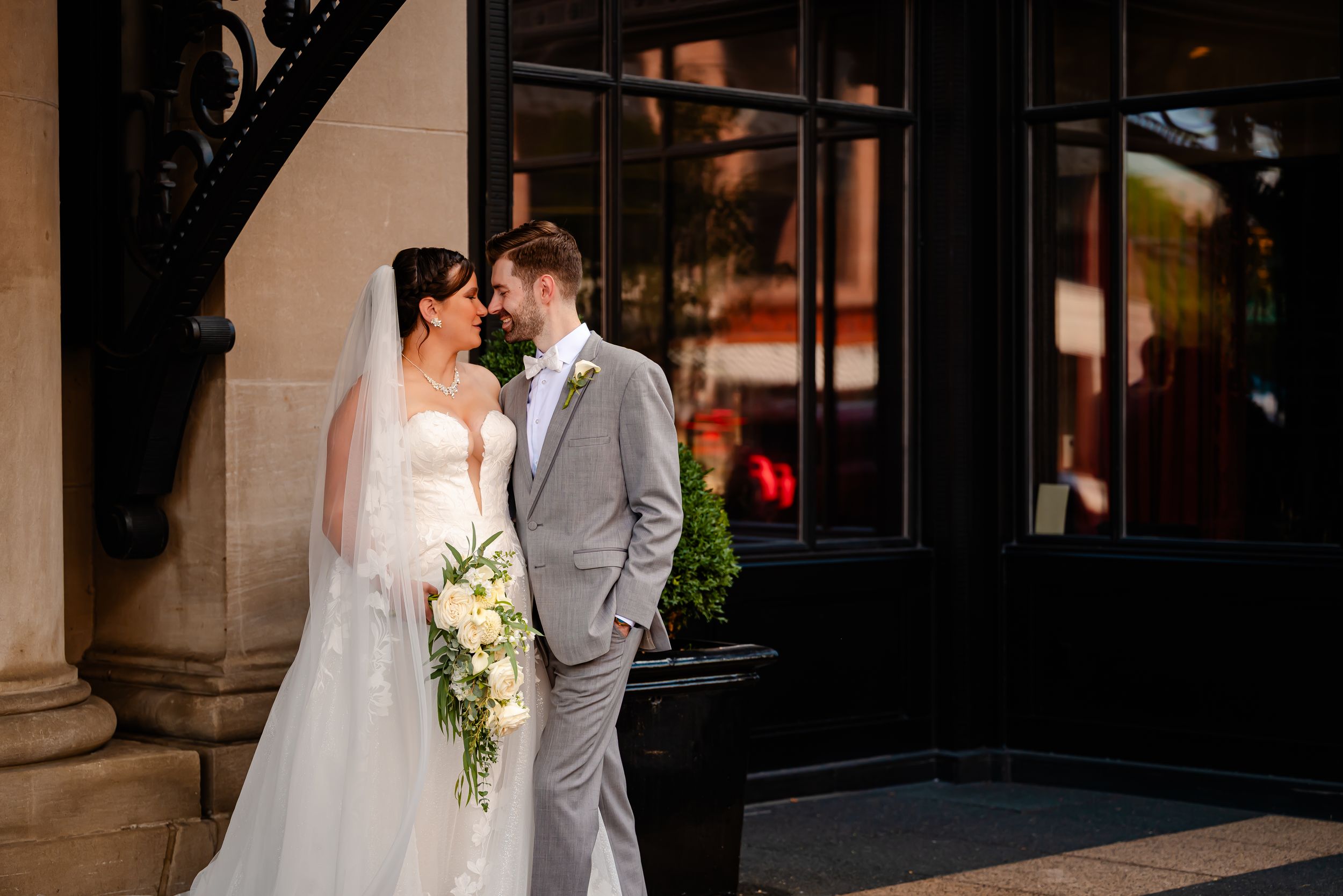 Bride and groom share a romantic moment outside a elegant venue, bride holding white bouquet wearing veil and strapless gown.