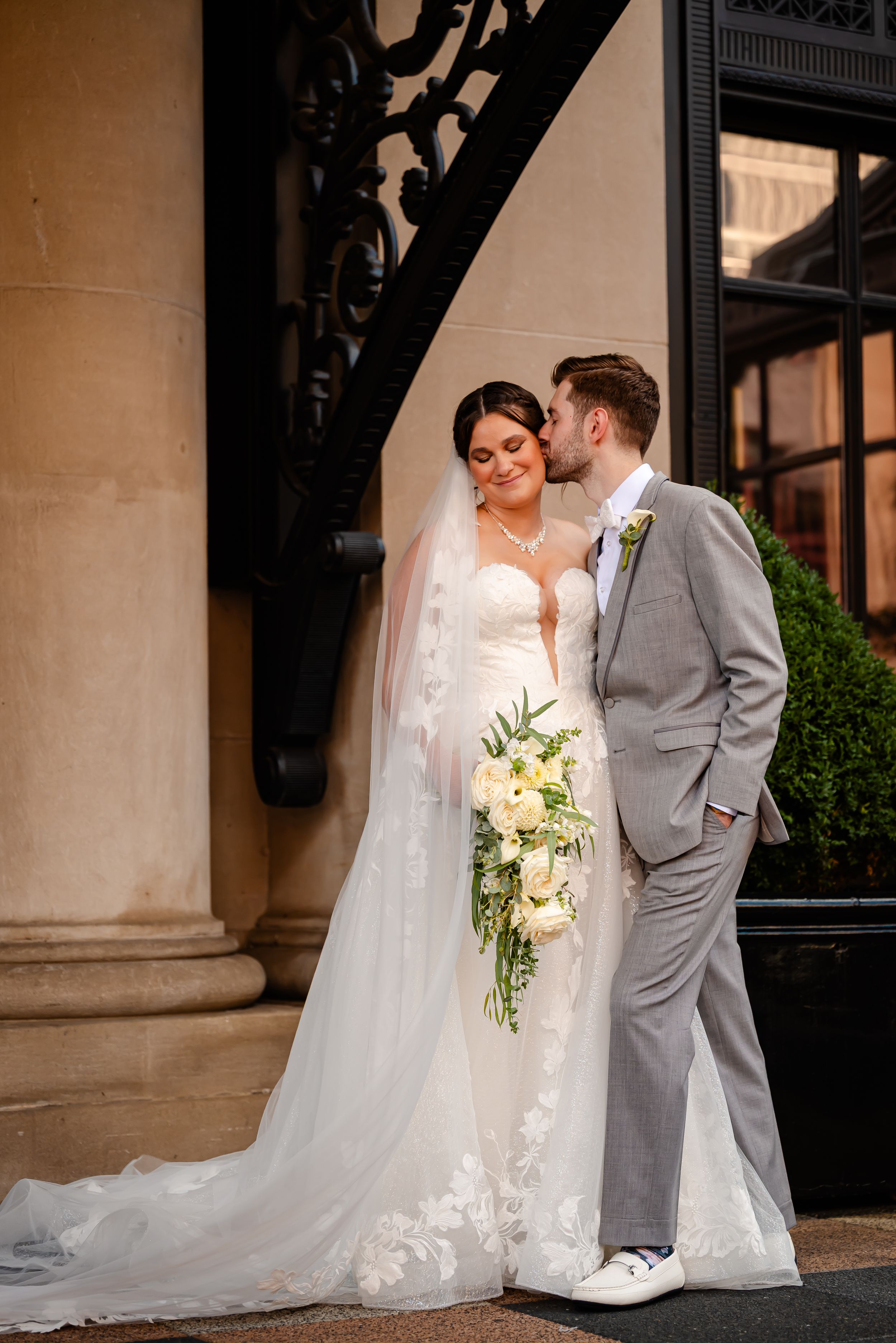 Bride in lace gown with veil holding white bouquet, groom in gray suit kissing her cheek outside elegant venue.