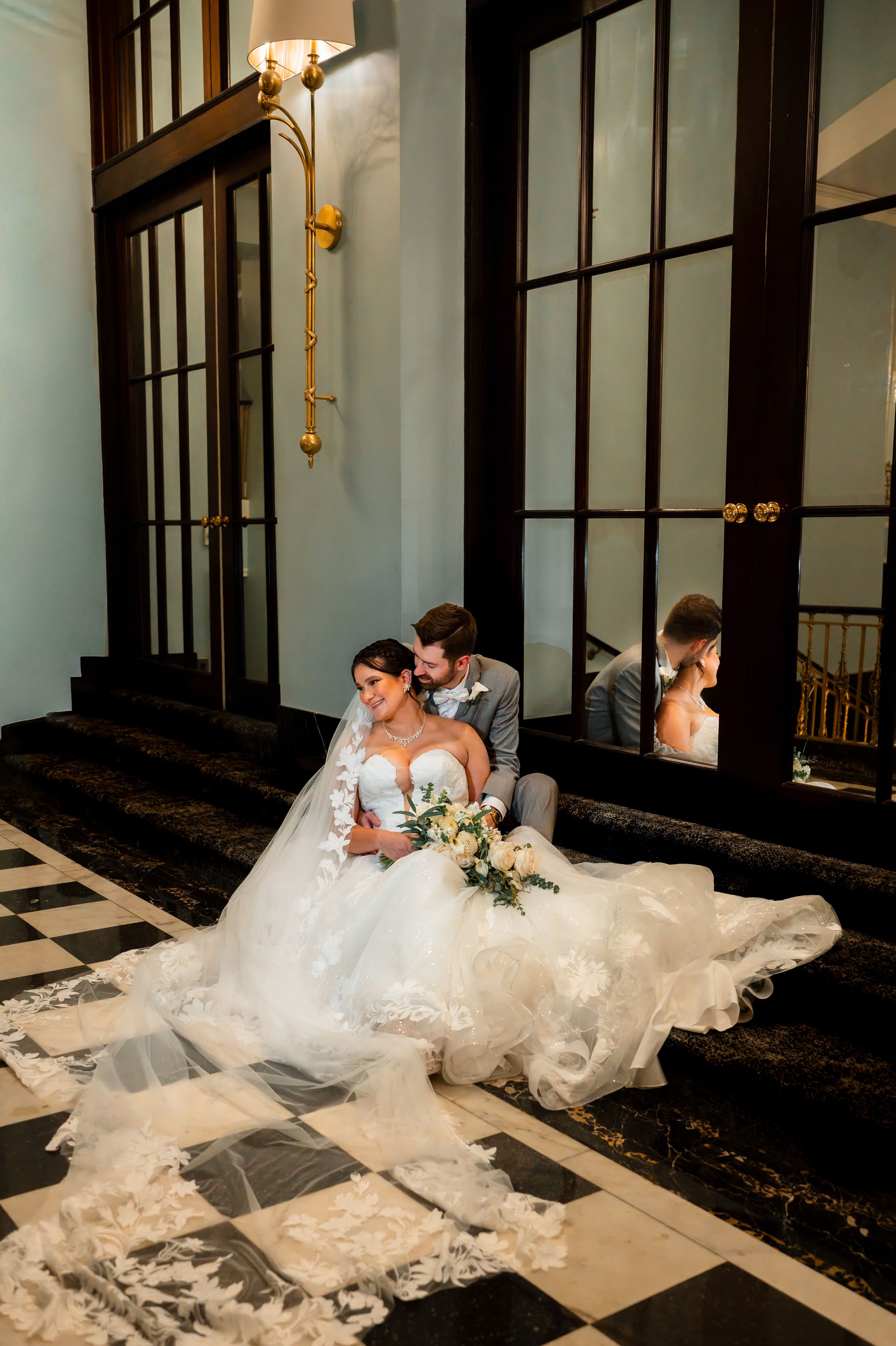 Bride in lace ballgown with veil sits on marble steps with groom, bouquet in hand, reflected in elegant French doors.