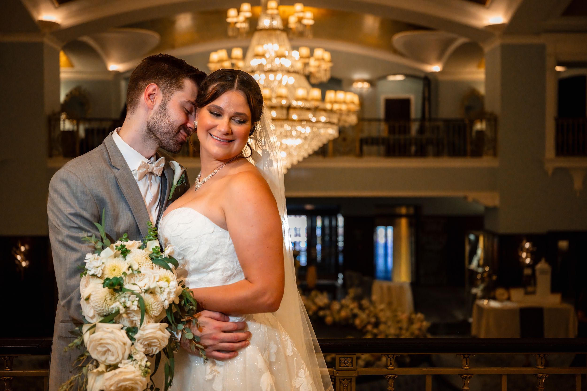 Bride and groom embrace elegantly under a golden chandelier in a grand ballroom, bride holding white rose bouquet.