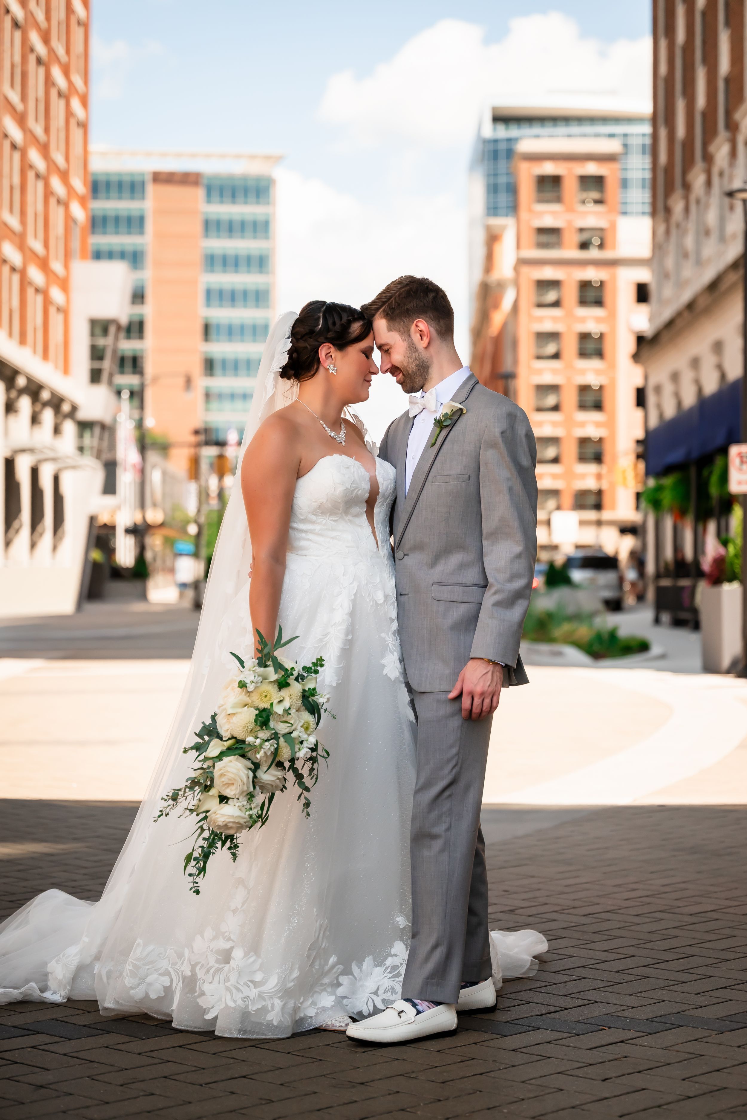 Bride and groom share romantic nose-to-nose moment in urban setting, bride holding white floral bouquet in lace wedding gown.
