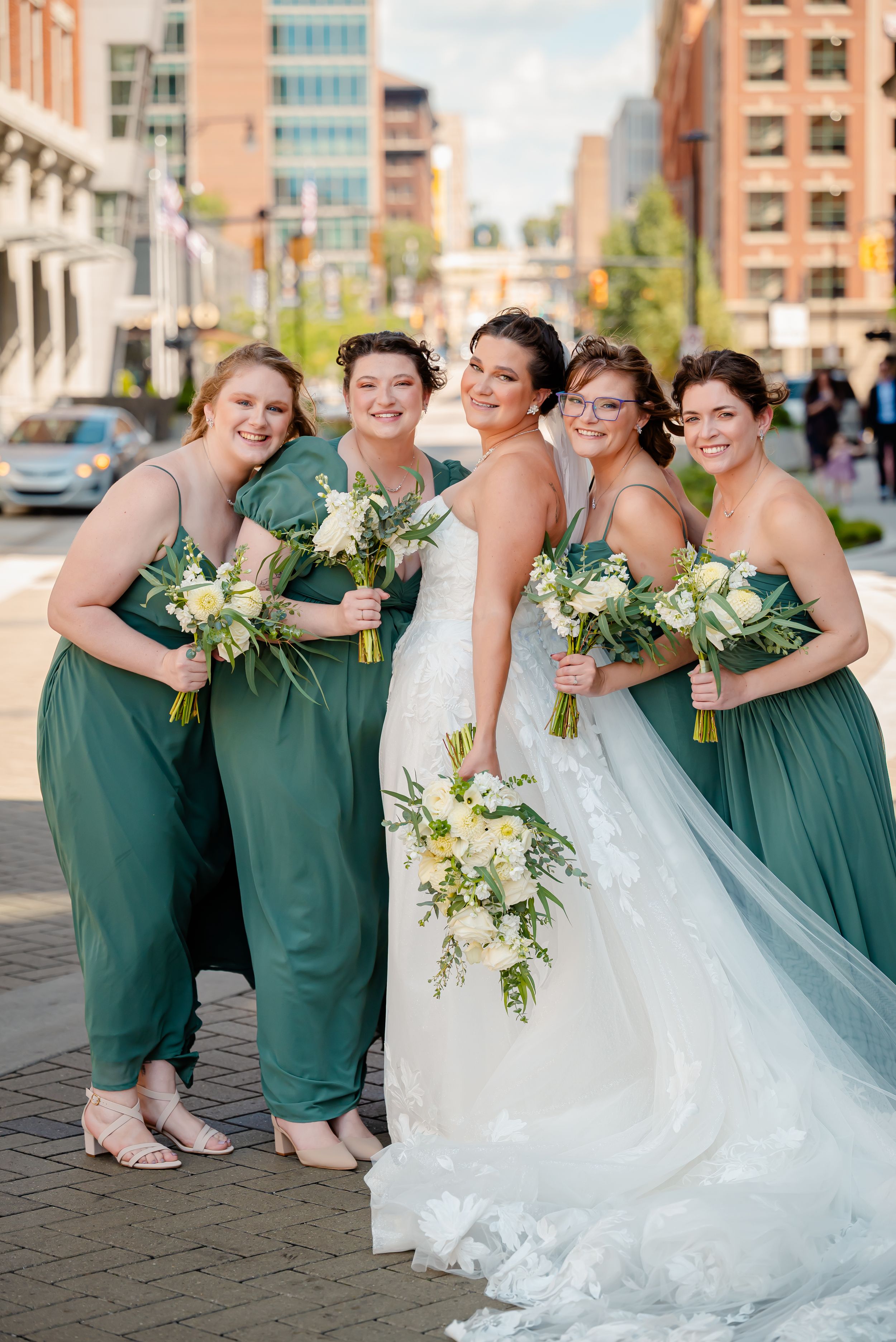 Bride in white gown poses with four bridesmaids in green dresses holding white bouquets on a city street.