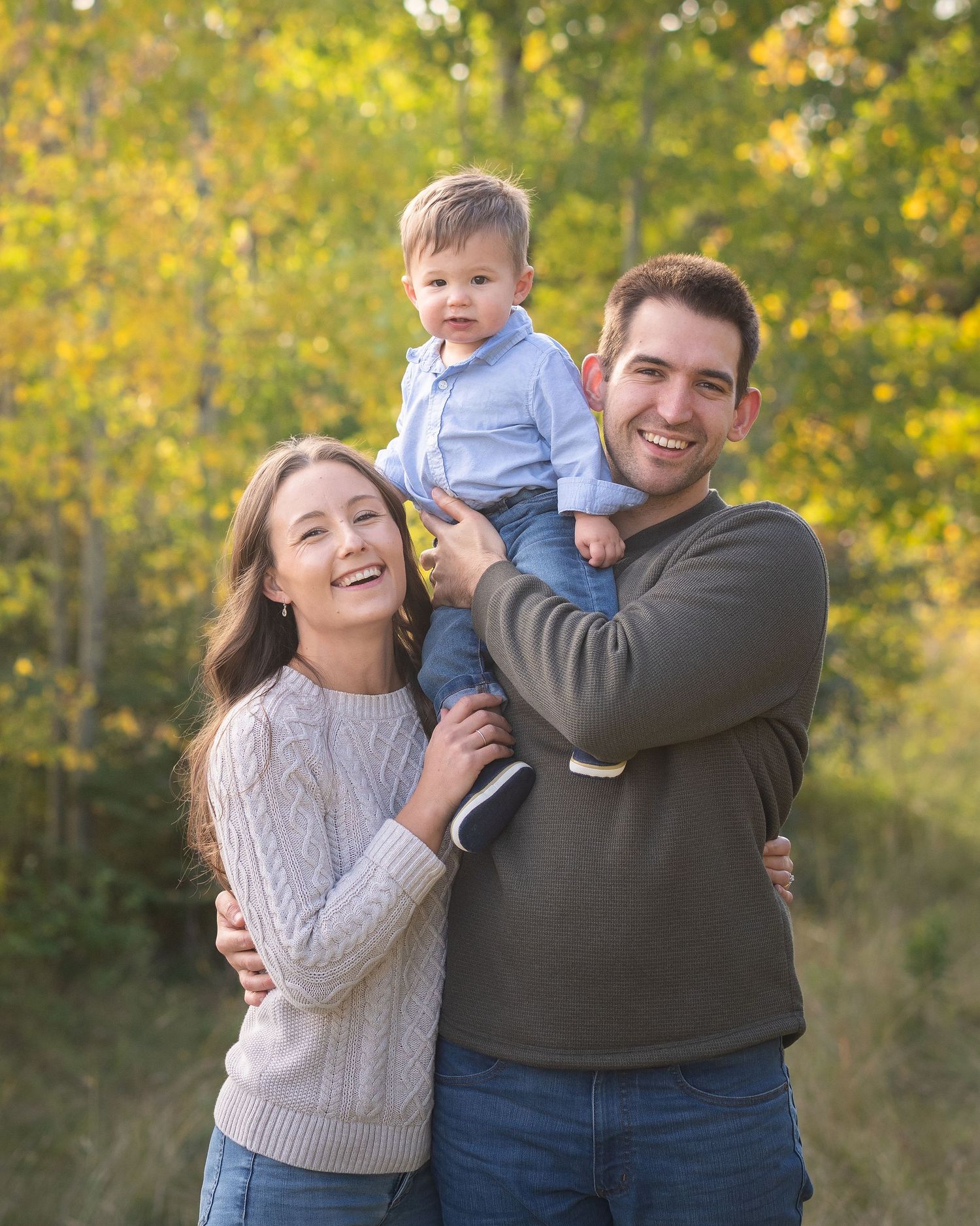 A smiling family of three poses outdoors in fall foliage, with a man holding a toddler boy beside a woman in a white sweater.