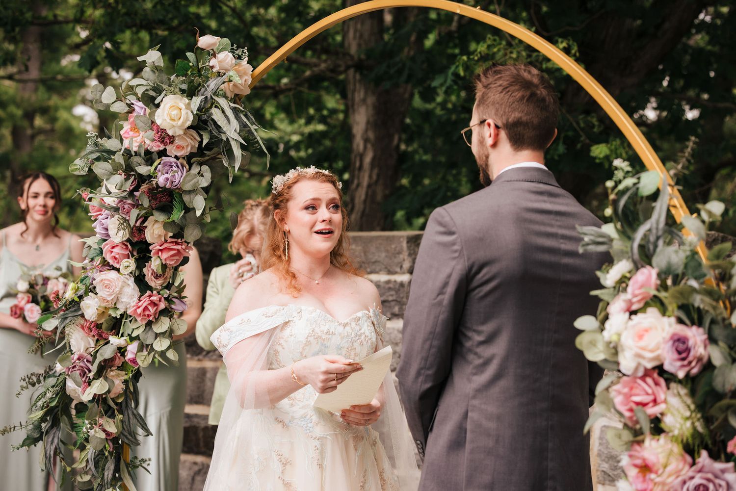 Bride reading handwritten vows to the groom under a gold arch with pink and purple florals at Searles Castle in Windham, New Hampshire.