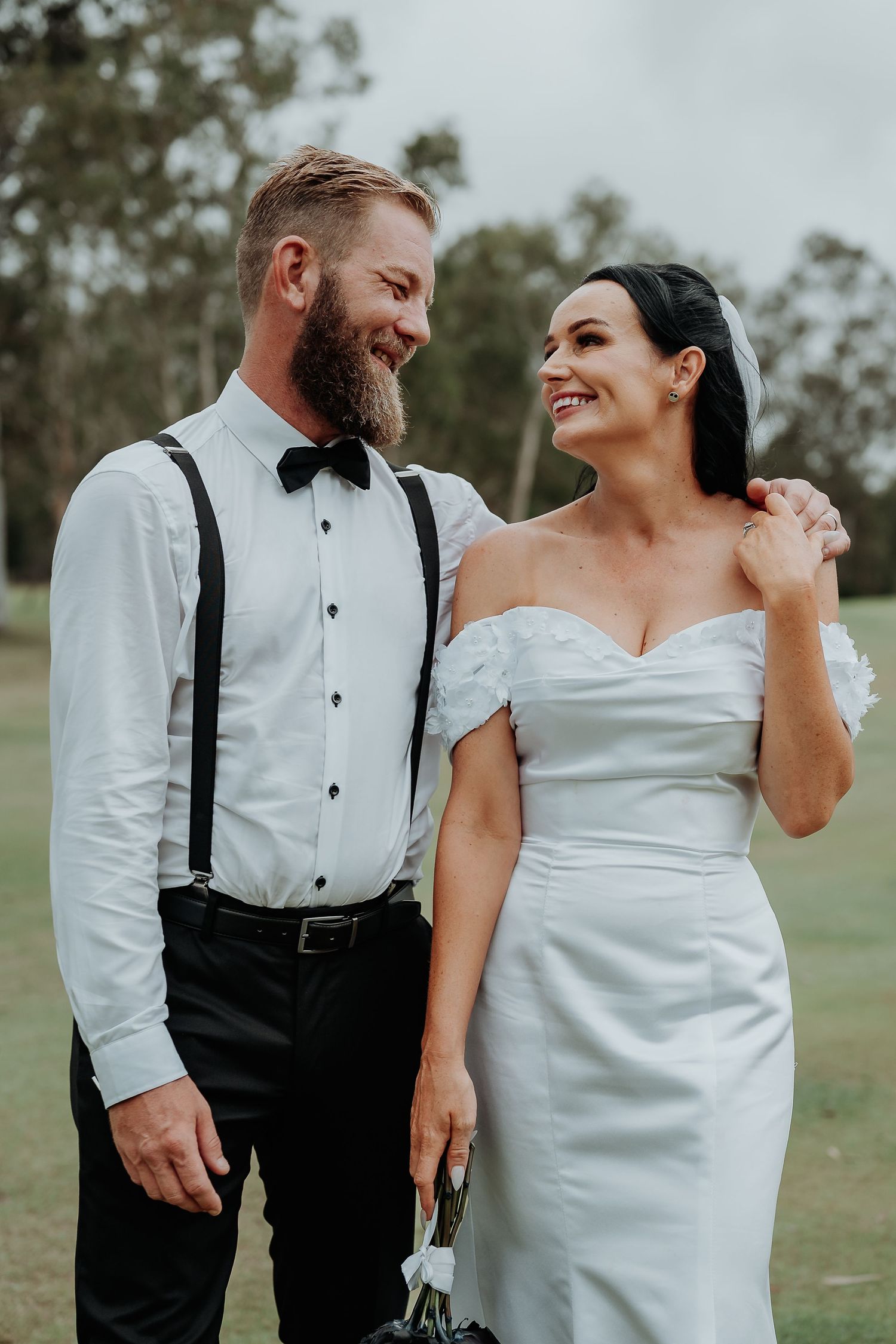 Smiling bride in white off-shoulder gown and groom in suspenders and bow tie share a joyful moment outdoors.