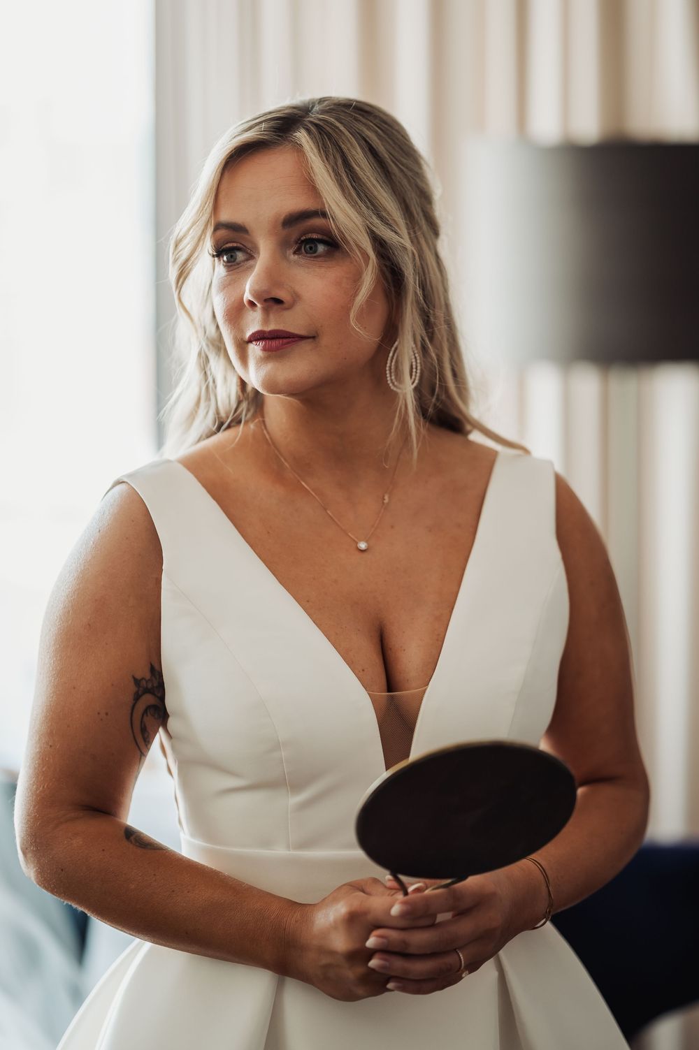 Bride in white deep V-neck gown holding a mirror, with elegant jewelry and soft wavy hair.