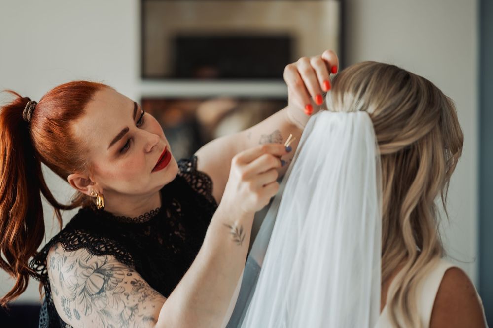 Red-haired tattoo artist attaching a bridal veil to a blonde bride's hair during wedding preparation.